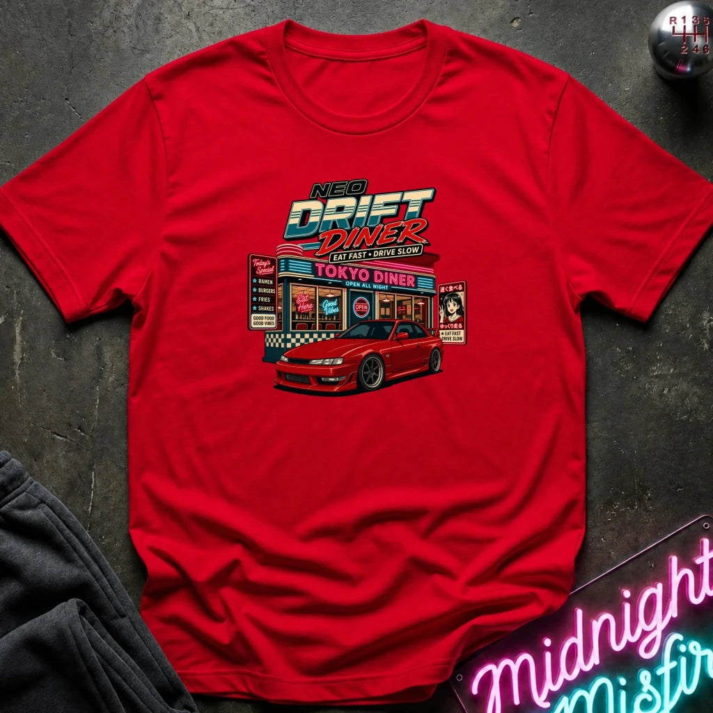 A red short-sleeve graphic t-shirt laid flat on a dark textured surface, featuring a colorful 'Nex Drift Diner' Tokyo Drift car diner illustration on the front. A pink neon sign and a dark sweatshirt are partially visible as props.