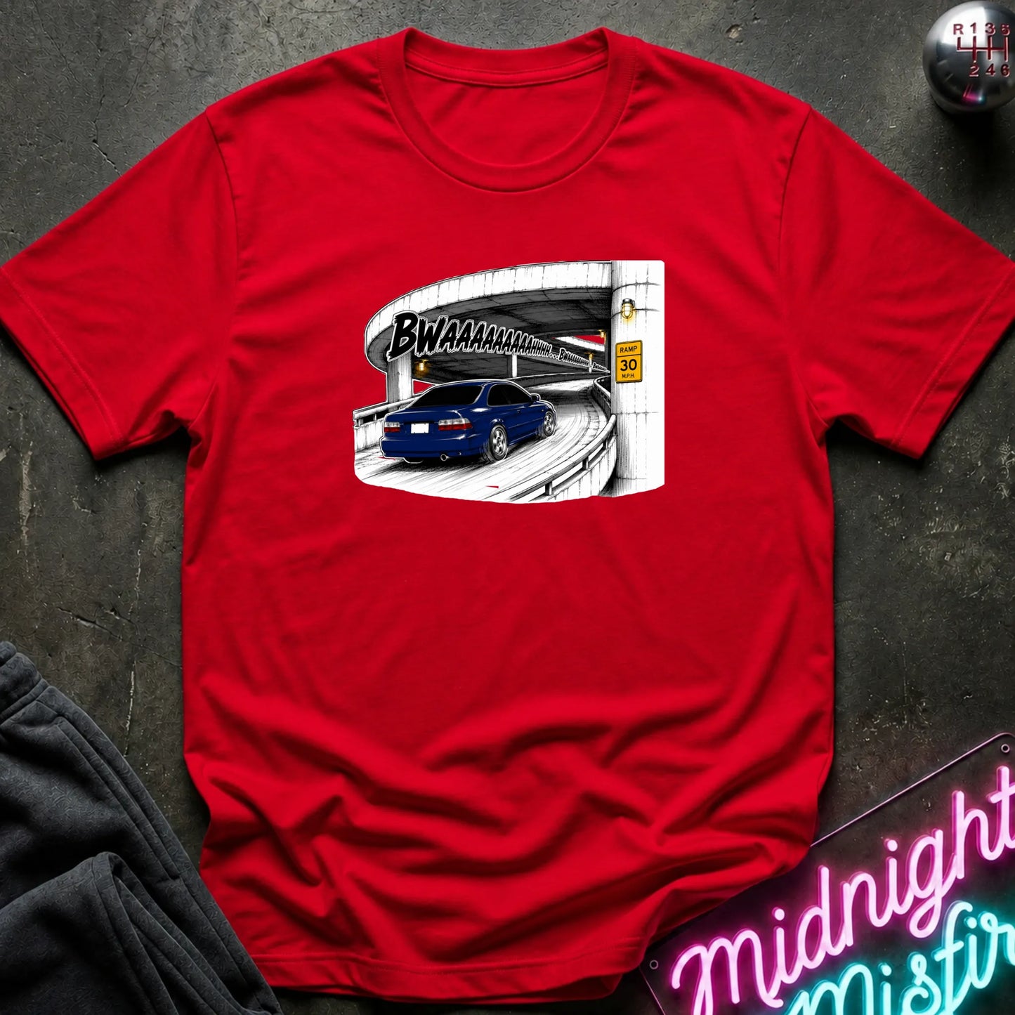 A red short-sleeve crewneck t-shirt laid flat on a dark textured surface. The shirt features a graphic print of a blue car driving under a highway overpass with the text 'BWAHAHA'.