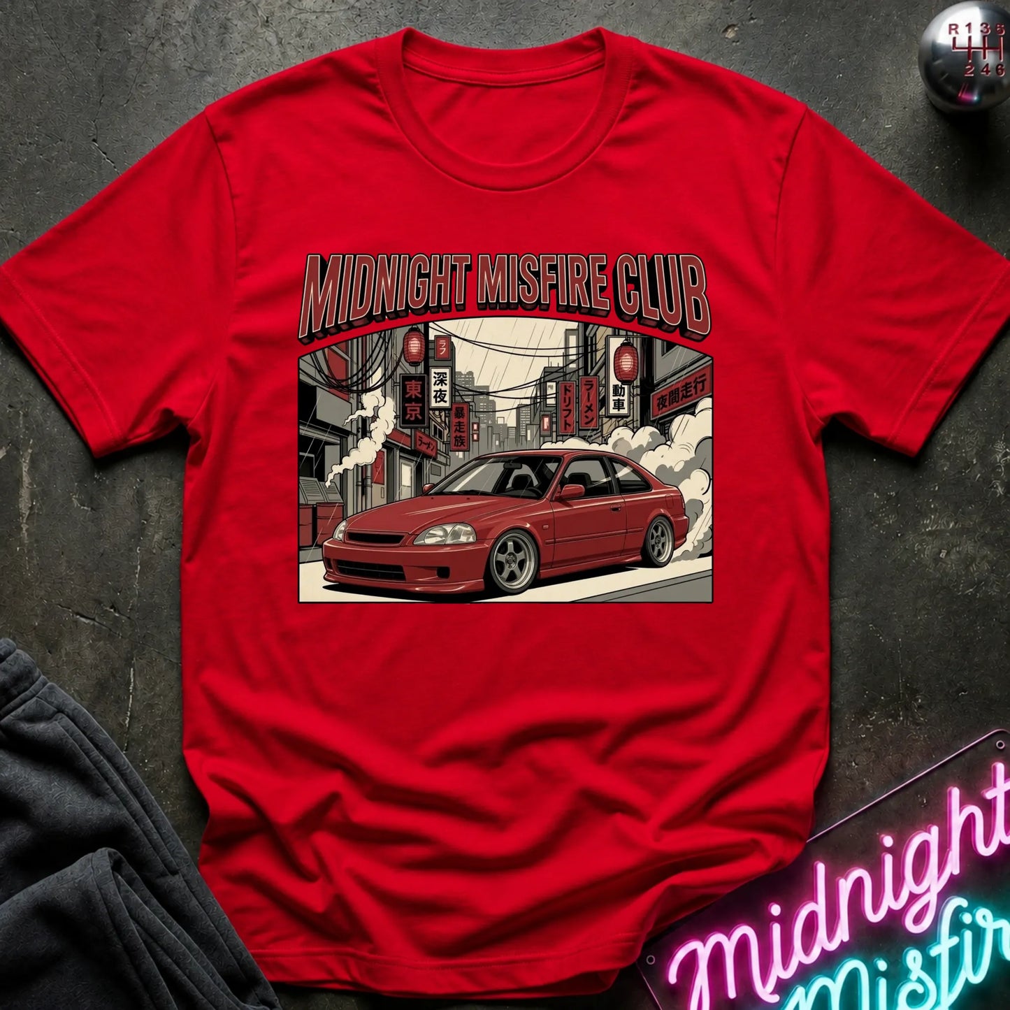 A red short-sleeve graphic t-shirt laid flat on a dark textured surface. The shirt features a central illustration of a red coupe car in a city street scene with the text 'MIDNIGHT MISFIRE CLUB' above it.