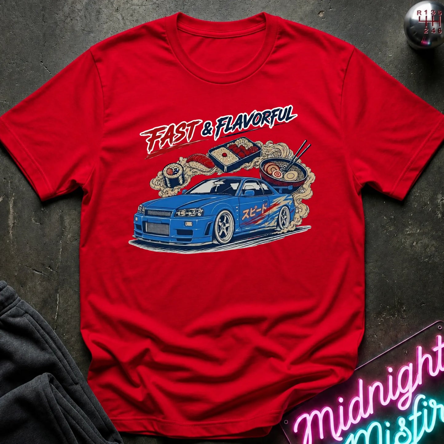 A red short-sleeve graphic t-shirt laid flat on a dark textured surface. The shirt features a colorful graphic of a blue sports car with smoke, sushi, and the text 'FAST & FLAVORFUL' in stylized lettering.