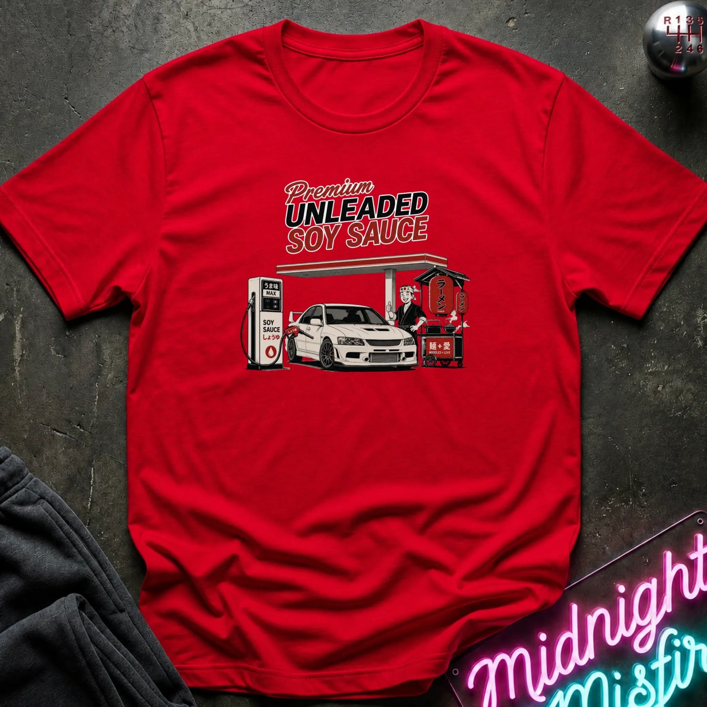A red short-sleeve graphic t-shirt laid flat on a dark textured surface. The graphic features the words 'Premium UNLEADED SOY SAUCE' above an illustration of a white car at a gas station pump.