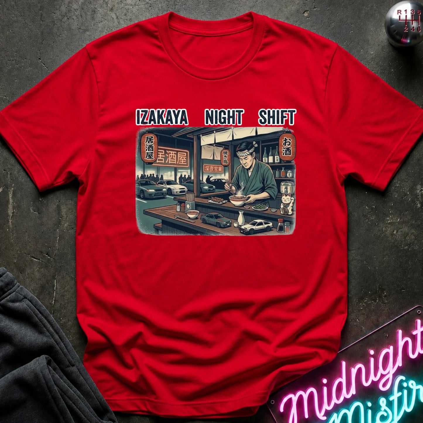 A red short-sleeve t-shirt laid flat on a dark textured surface, featuring a vintage-style graphic of an izakaya scene with the text 'IZAKAYA NIGHT SHIFT' above it. A neon sign and dark clothing are partially visible in the styled flat-lay composition.