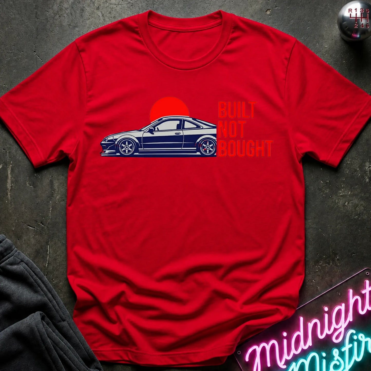 A red short-sleeve t-shirt laid flat on a dark textured surface, featuring a graphic of a blue sports coupe car in front of a red sun with the text 'BUILT NOT BOUGHT' in red. A neon sign and dark clothing are partially visible around the shirt.