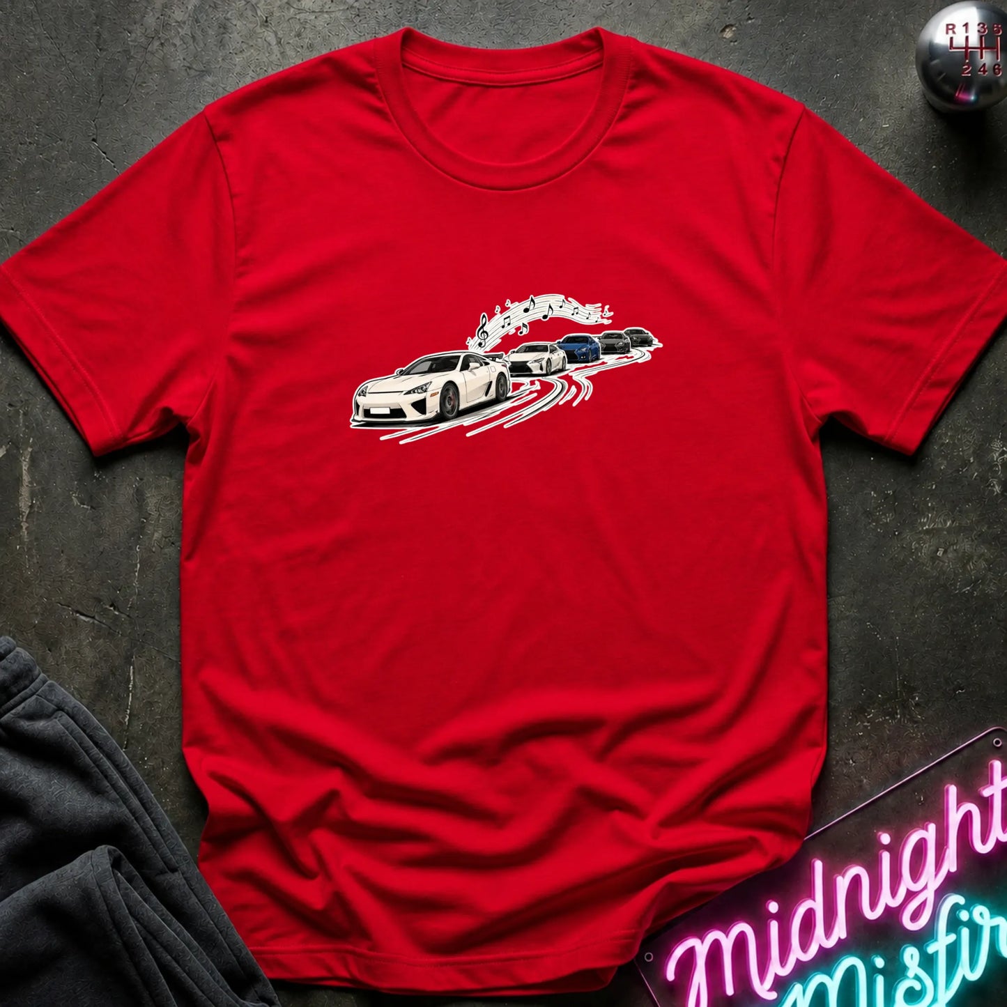 A bright red short-sleeve crewneck t-shirt featuring a graphic print of a white sports car leading a line of other cars, laid flat on a dark textured surface with a neon sign and dark pants partially visible.