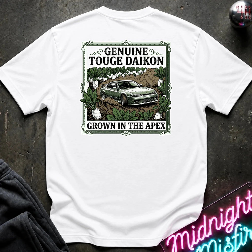 A white short-sleeve graphic T-shirt laid flat on a dark textured surface. The shirt features a rectangular vintage-style print with the words 'Genuine Touge Daikon' and 'Grown in the Apex' surrounding an illustration of a green car in a field.
