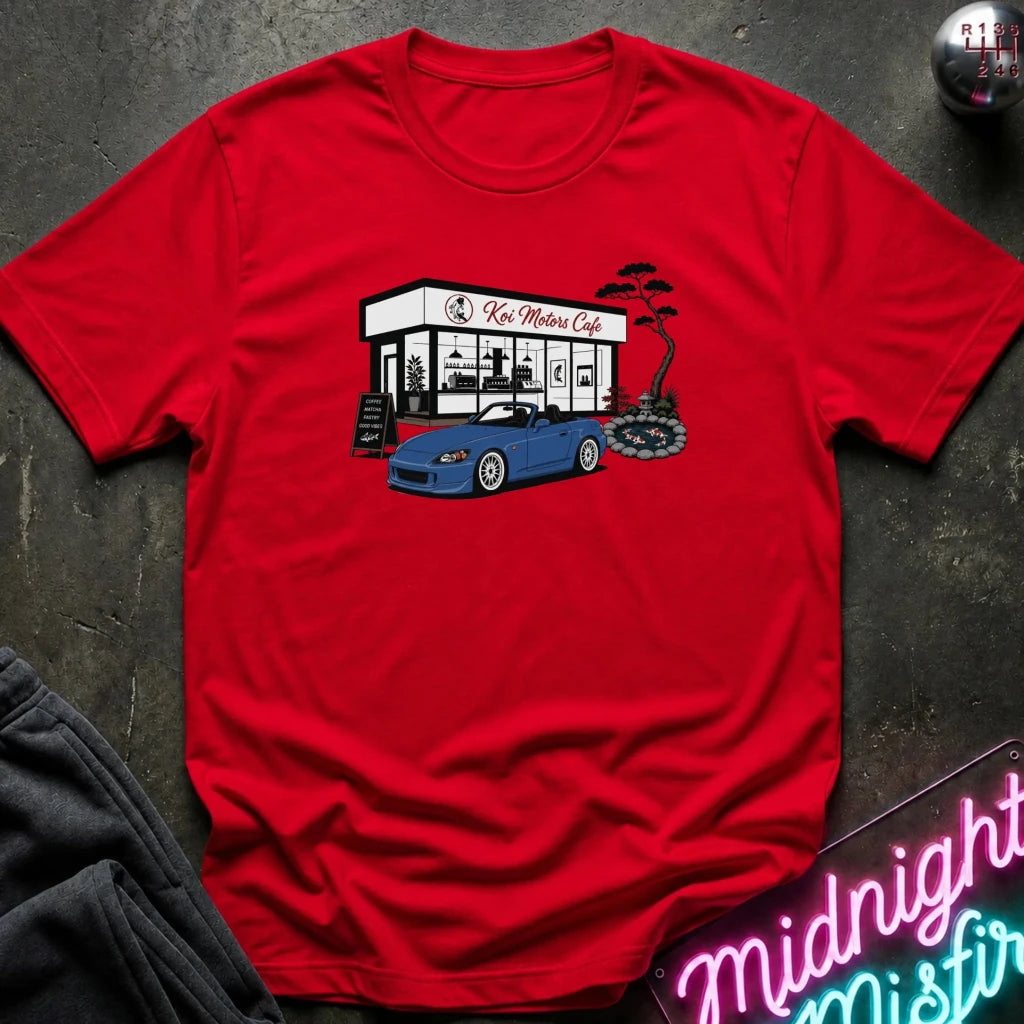 A red short-sleeve t-shirt laid flat on a dark textured surface, featuring a graphic print of a blue sports car parked in front of 'Koi Motors Cafe' with a small tree illustration. 
