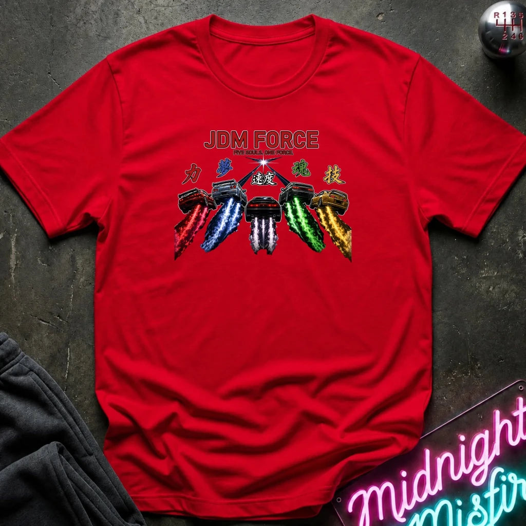 A red short-sleeve graphic T-shirt laid flat on a dark textured surface. The shirt features a colorful chest print with the text 'JDM FORCE' and stylized Japanese characters above five paint-splattered spray-paint cans in red, blue, white, green, and gold.