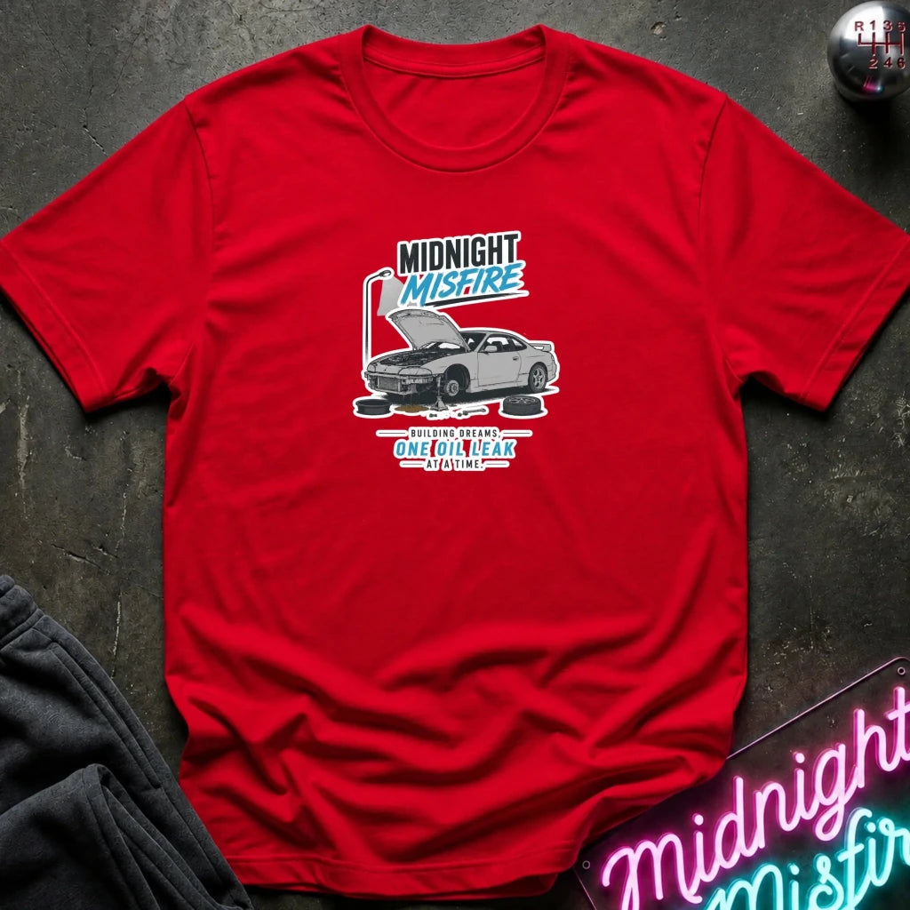 A red short-sleeve graphic T-shirt laid flat on a dark textured surface. The shirt features a central graphic of a gray sports car with its hood open, surrounded by text that reads 'MIDNIGHT MISFIRE'