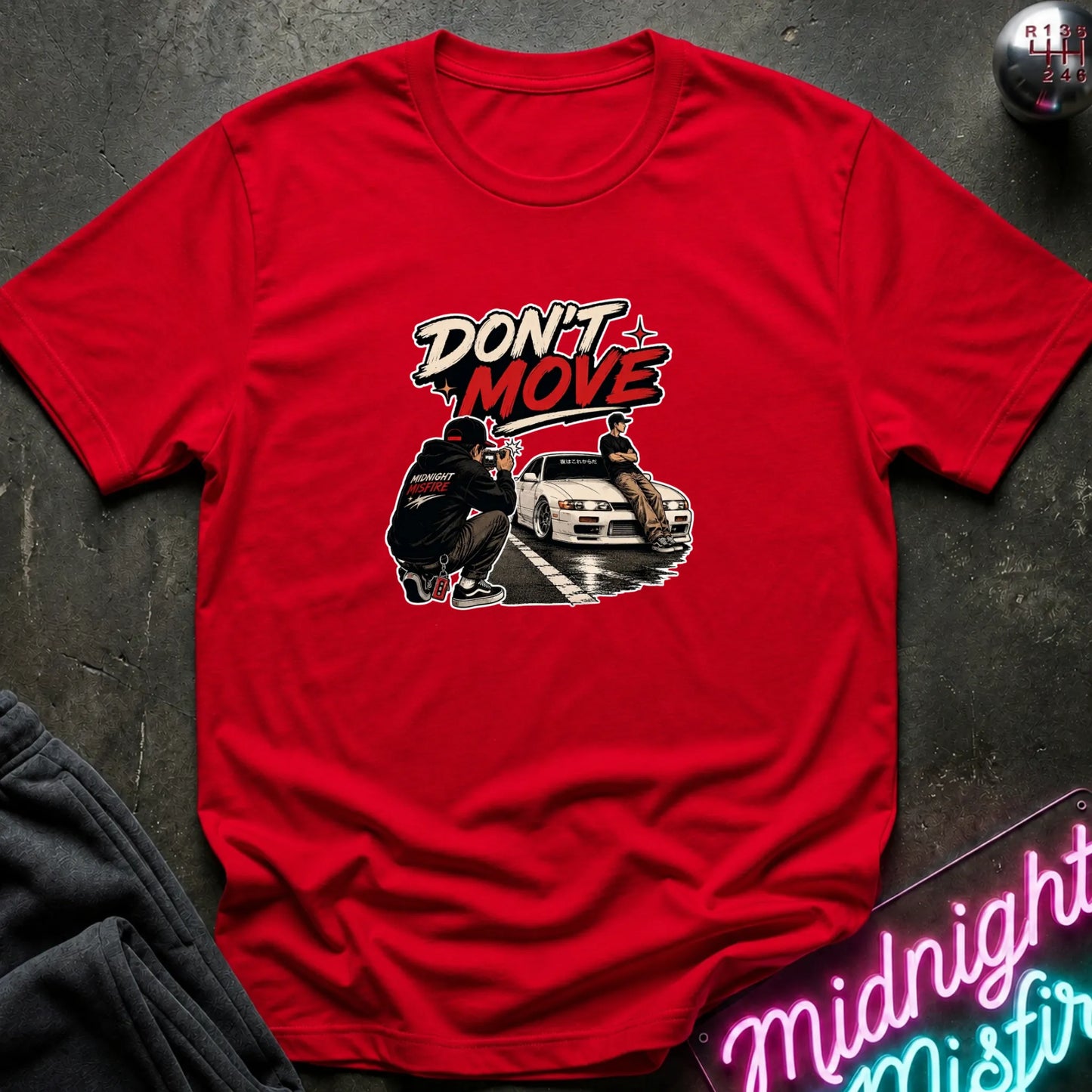 A red short-sleeve graphic t-shirt laid flat on a dark textured surface. The graphic features the words 'DON'T MOVE' in a stylized white and red font above an illustration of a person crouching next to a white sports car.