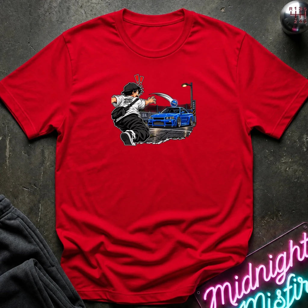 A red short-sleeve crewneck t-shirt laid flat on a dark textured surface, featuring a graphic print of a cartoon character next to a blue sports car. A gray sweatshirt is partially visible on the bottom left, and a neon sign and metallic ball are on the right.