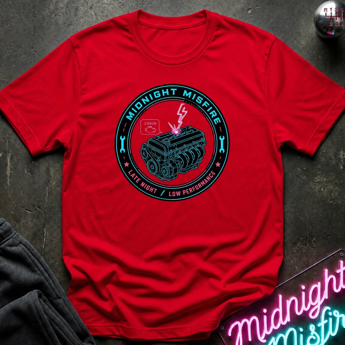 A red short-sleeve crewneck t-shirt laid flat on a dark textured surface. The shirt features a circular teal and black graphic in the center depicting a car engine with a lightning bolt and the text 'MIDNIGHT MISFIRE' and 'LATE NIGHT / LOW PERFORMANCE'.