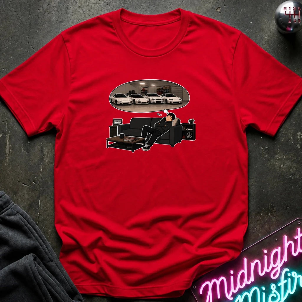 A red  short-sleeve graphic t-shirt laid flat on a dark textured surface. The graphic shows a man sitting on a black couch dreaming, with an oval reflection of several white cars above him.