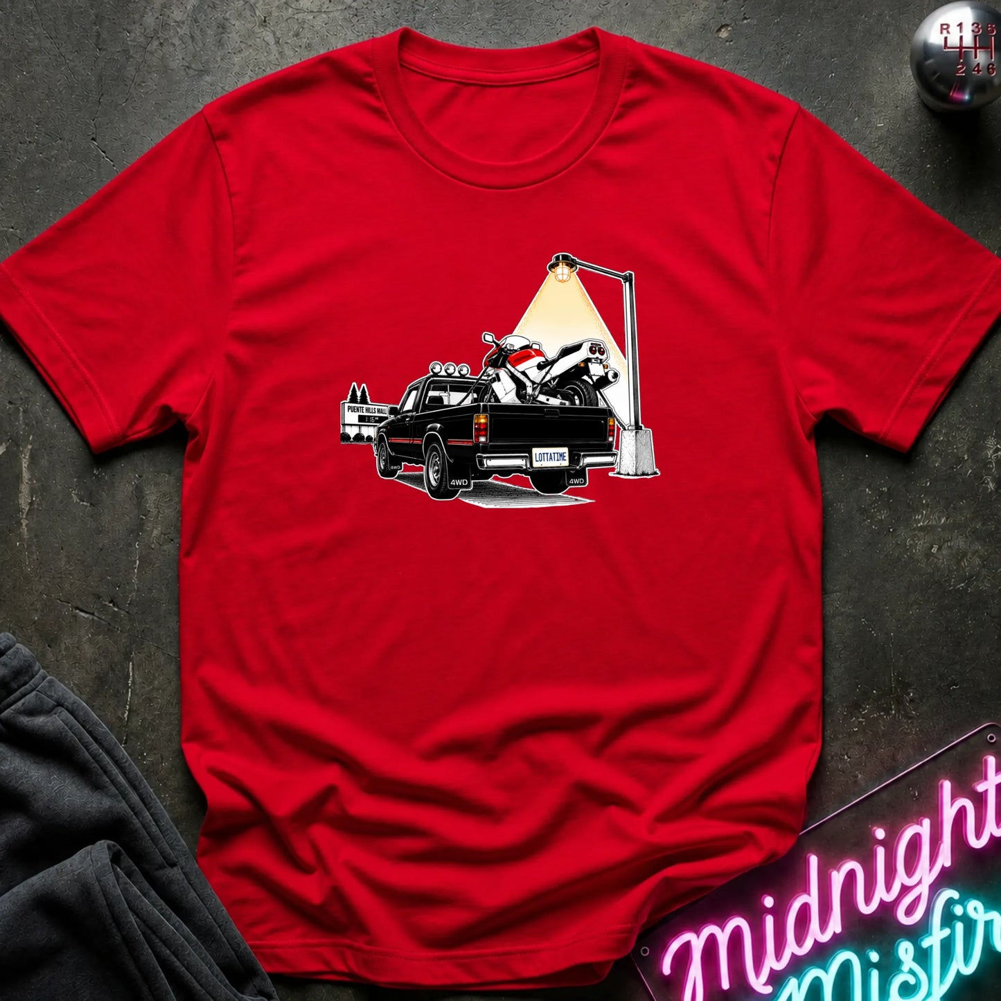 A red short-sleeve crewneck t-shirt featuring a graphic print of a black pickup truck carrying a white and red motorcycle under a street lamp, laid flat on a dark textured surface.