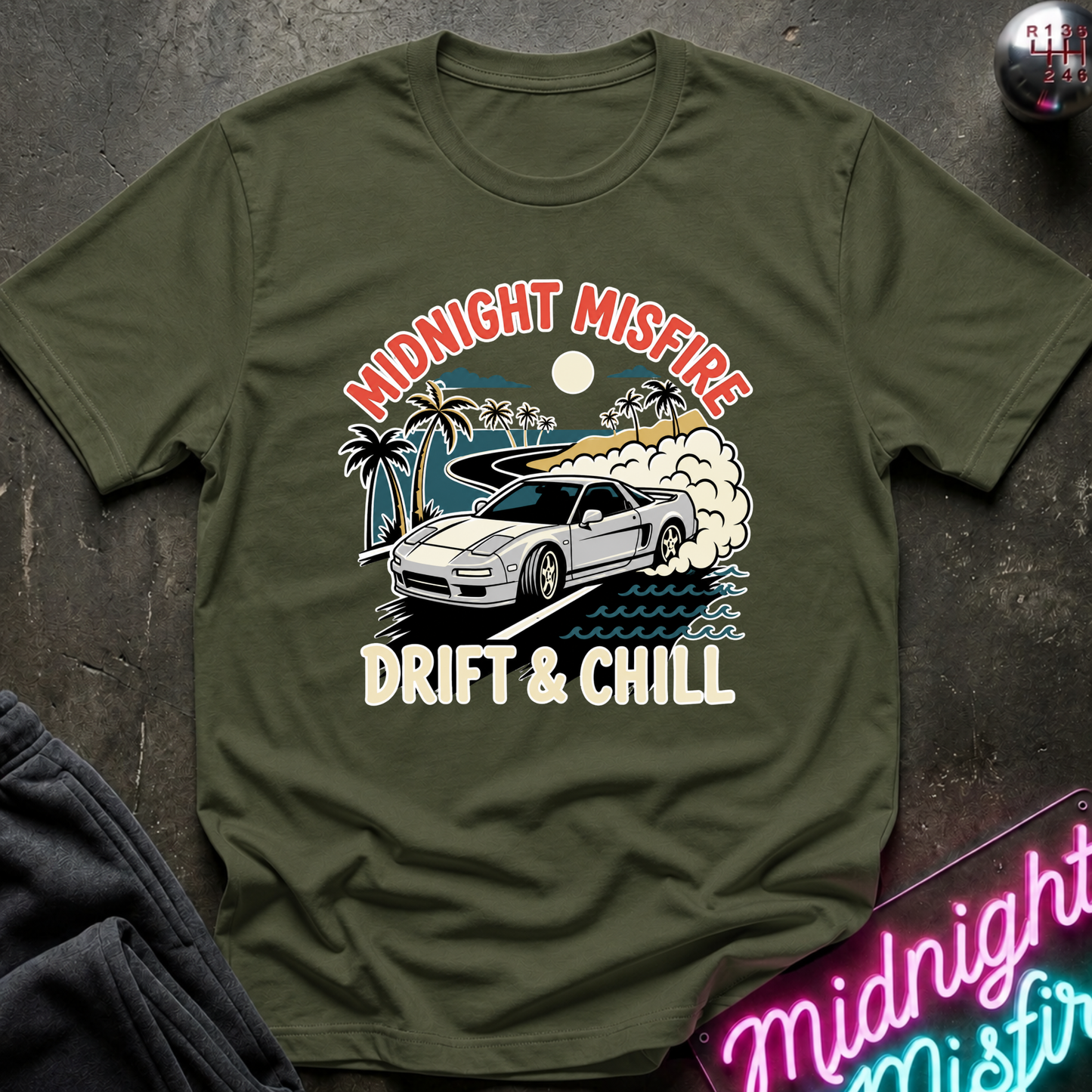 An olive green short-sleeve t-shirt with a graphic print of a white drift car, palm trees, and a sunset, featuring the text 'MIDNIGHT MISFIRE' and 'DRIFT & CHILL'. The shirt is laid flat on a dark textured surface with a neon sign and dark fabric in the corners.