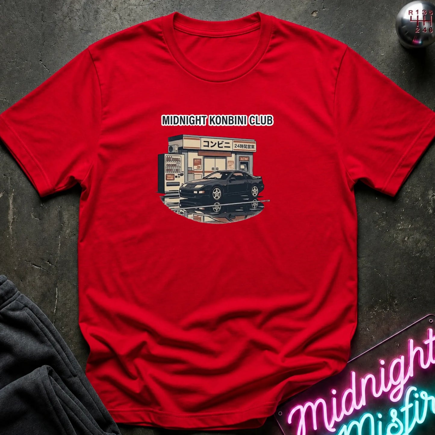 A red short-sleeve graphic t-shirt laid flat on a dark textured surface. The graphic shows a black sports car parked in front of a convenience store with the text 'MIDNIGHT KONBINI CLUB' above it.