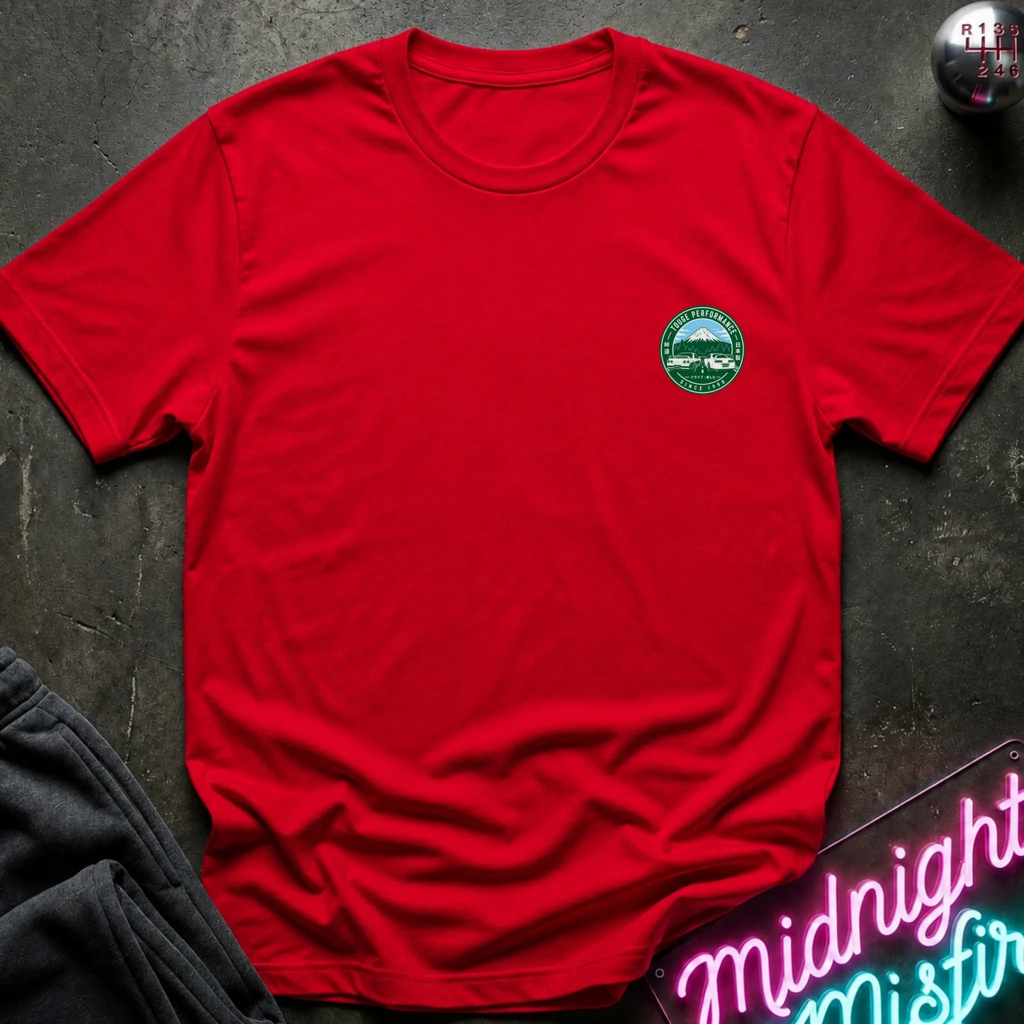 Red short-sleeve graphic T-shirt laid flat on a dark textured surface. The shirt features a small circular green and white print logo  with a mountain, two cars, and the text 'TOUGE PERFORMANCE' and 'SINCE 1990'.