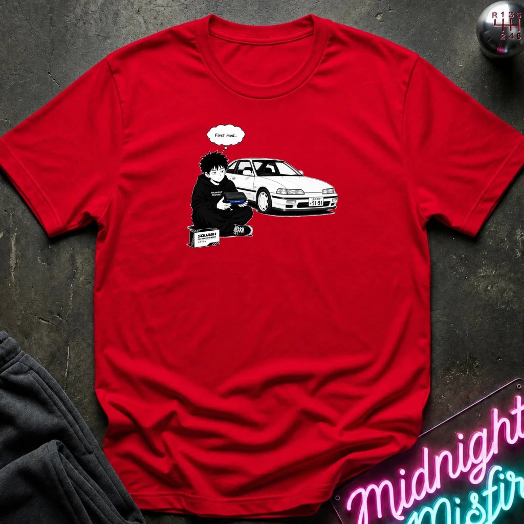 A red short-sleeve crewneck t-shirt laid flat on a dark textured surface. The shirt features a graphic print of a person in black sitting next to a white sports car with a speech bubble 