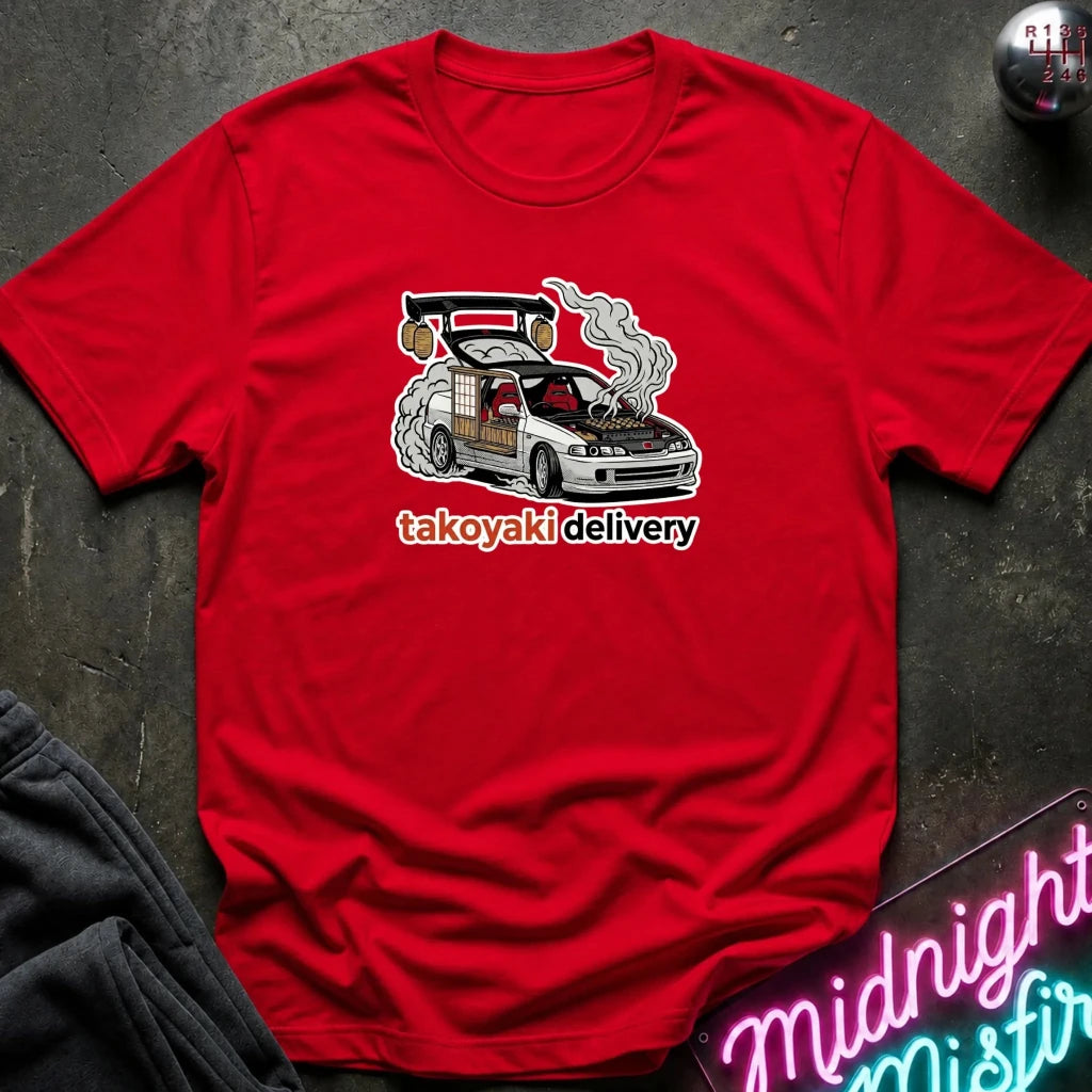 A red short-sleeve crewneck t-shirt laid flat on a dark textured surface. The shirt features a graphic of a white car with a large rear spoiler, smoke, and a basketball, with the text 'takoyaki delivery' printed below in white and orange.