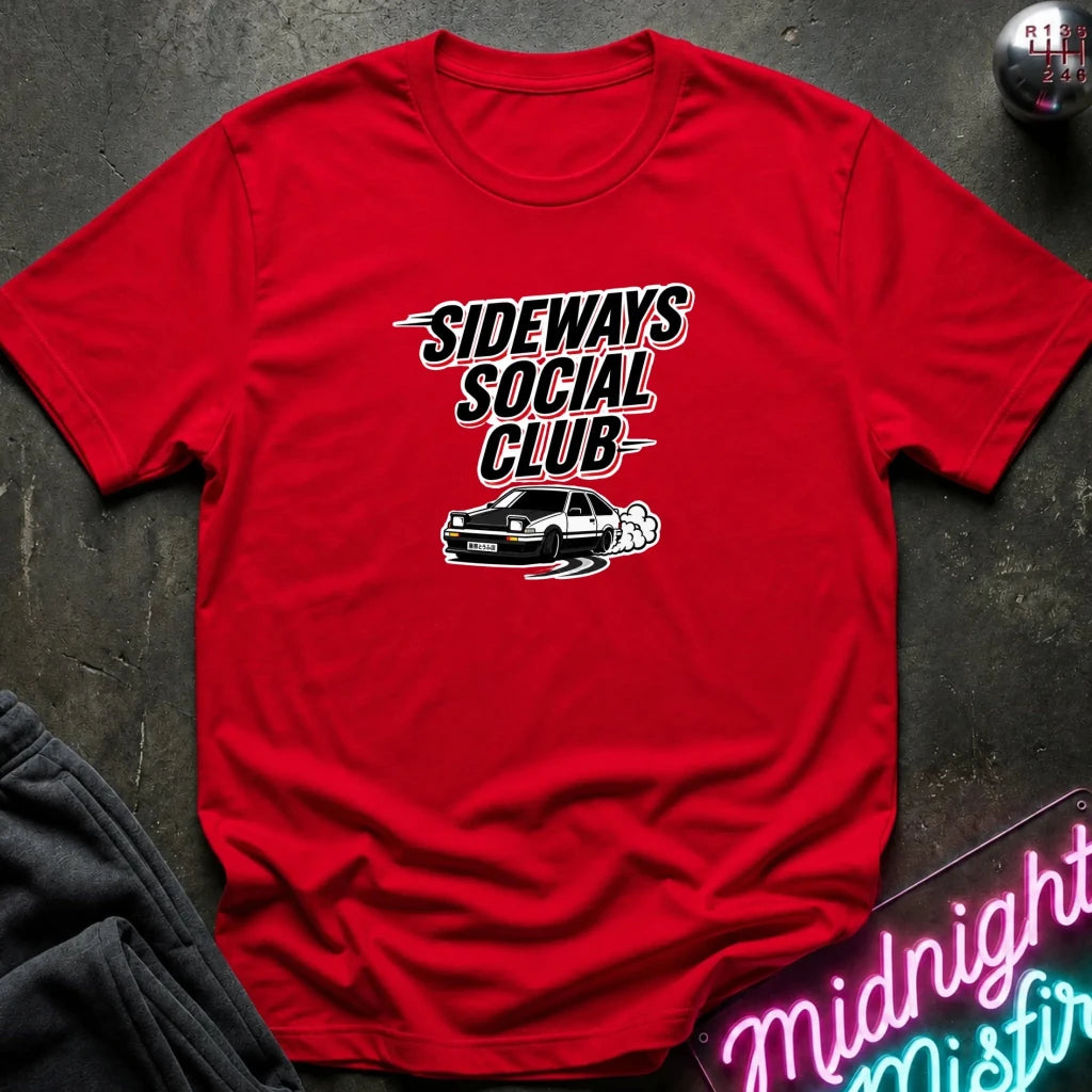 A red short-sleeve graphic T-shirt laid flat on a dark textured surface, featuring the words 'SIDEWAYS SOCIAL CLUB' in white and black with a small car illustration underneath. A gray sweatshirt corner and a neon-style sign are partially visible as styling props.