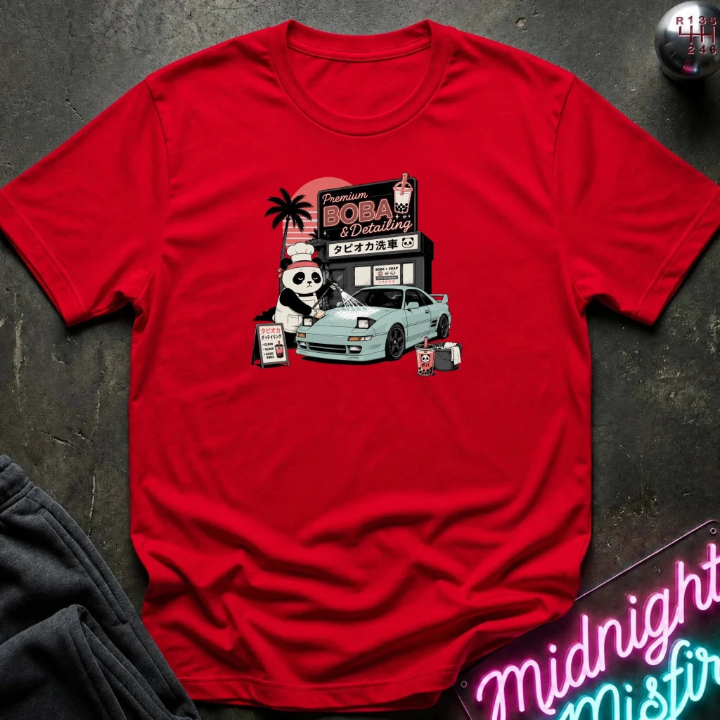 red short-sleeve graphic t-shirt laid flat on a dark textured surface. The graphic features a cartoon panda washinga light blue car