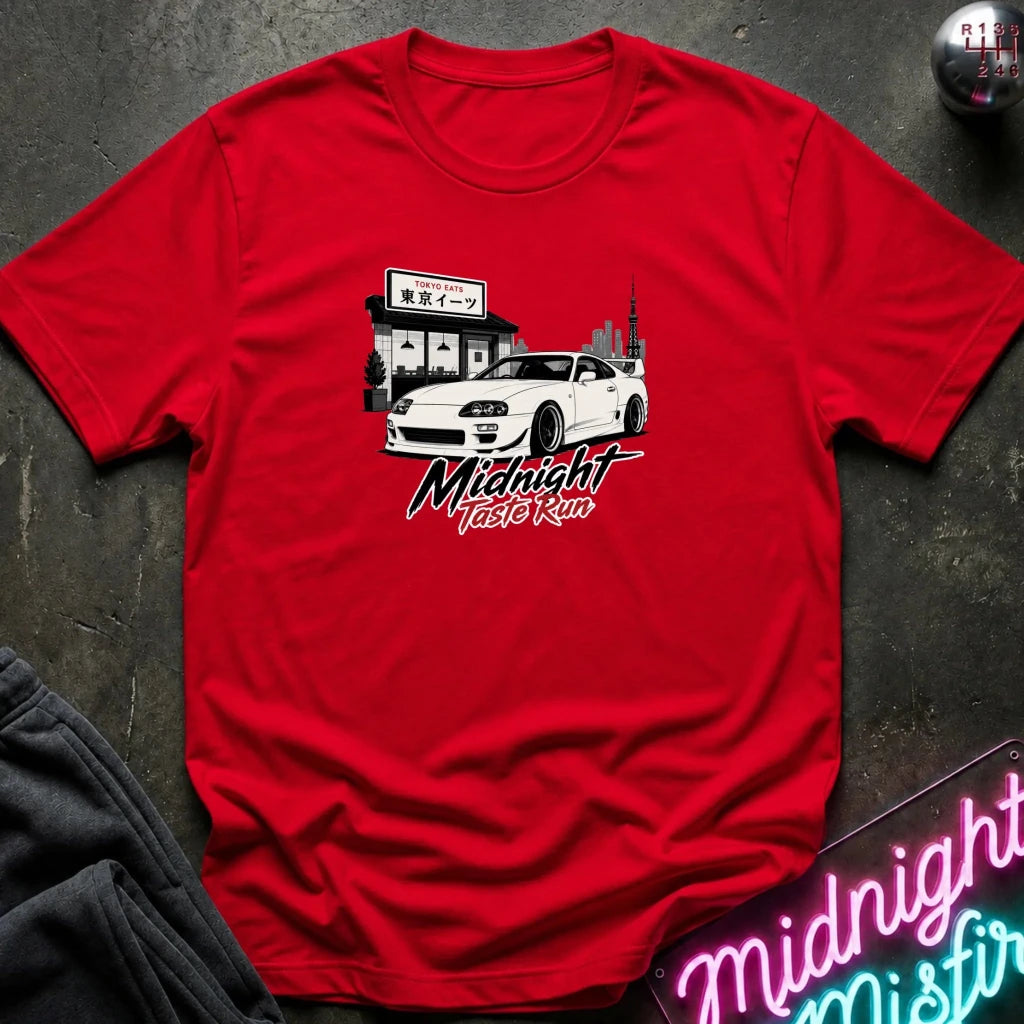 A red short-sleeve crewneck t-shirt laid flat on a dark textured surface. The shirt features a graphic print of a white sports car parked in front of a building with Japanese signage, and stylized text reading 'Midnight Taste Run' below.