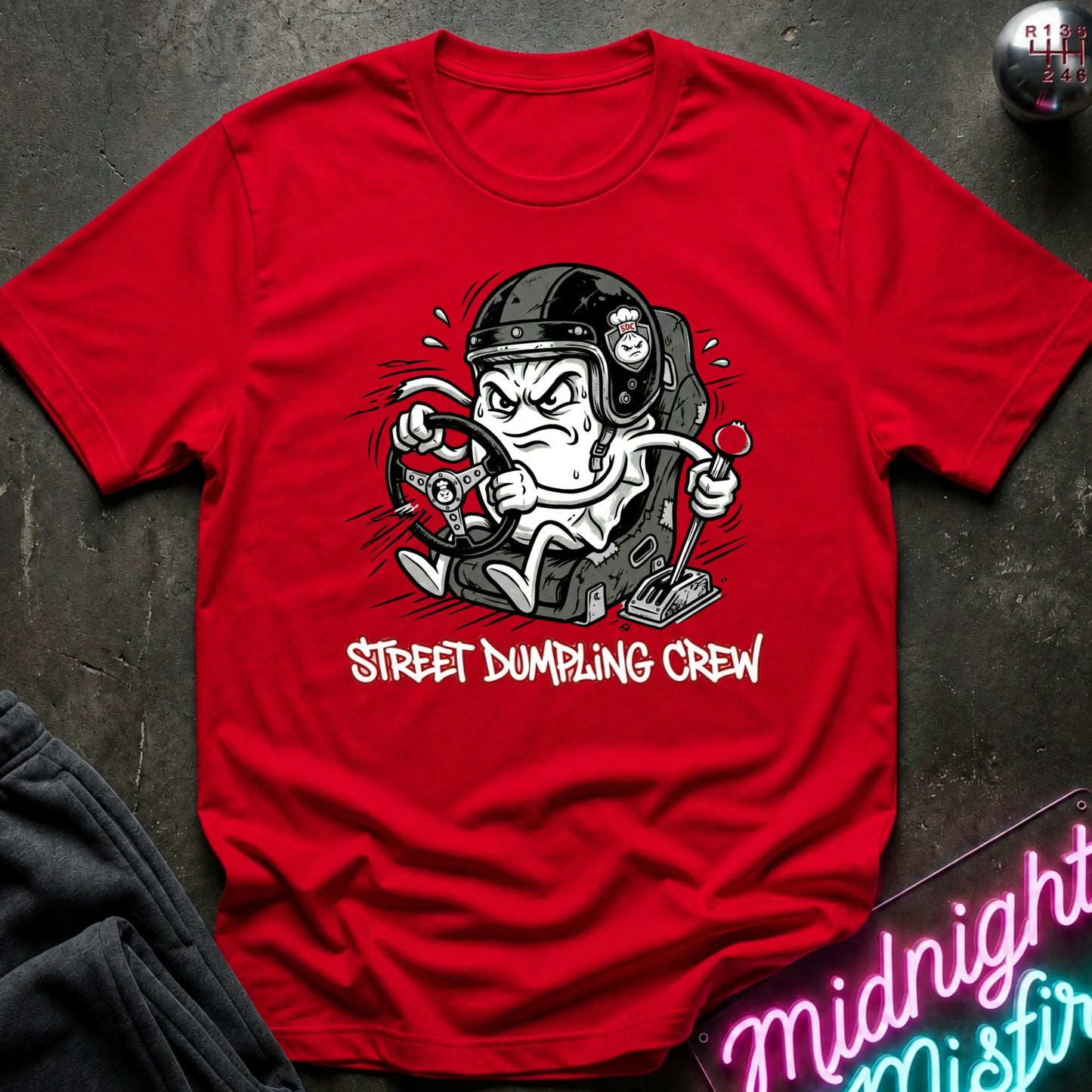 A red graphic t-shirt laid flat on a dark textured surface. The shirt features a cartoon illustration of a street-dumping character in a helmet with the text 'STREET DUMPING CREW' printed below in red and white.