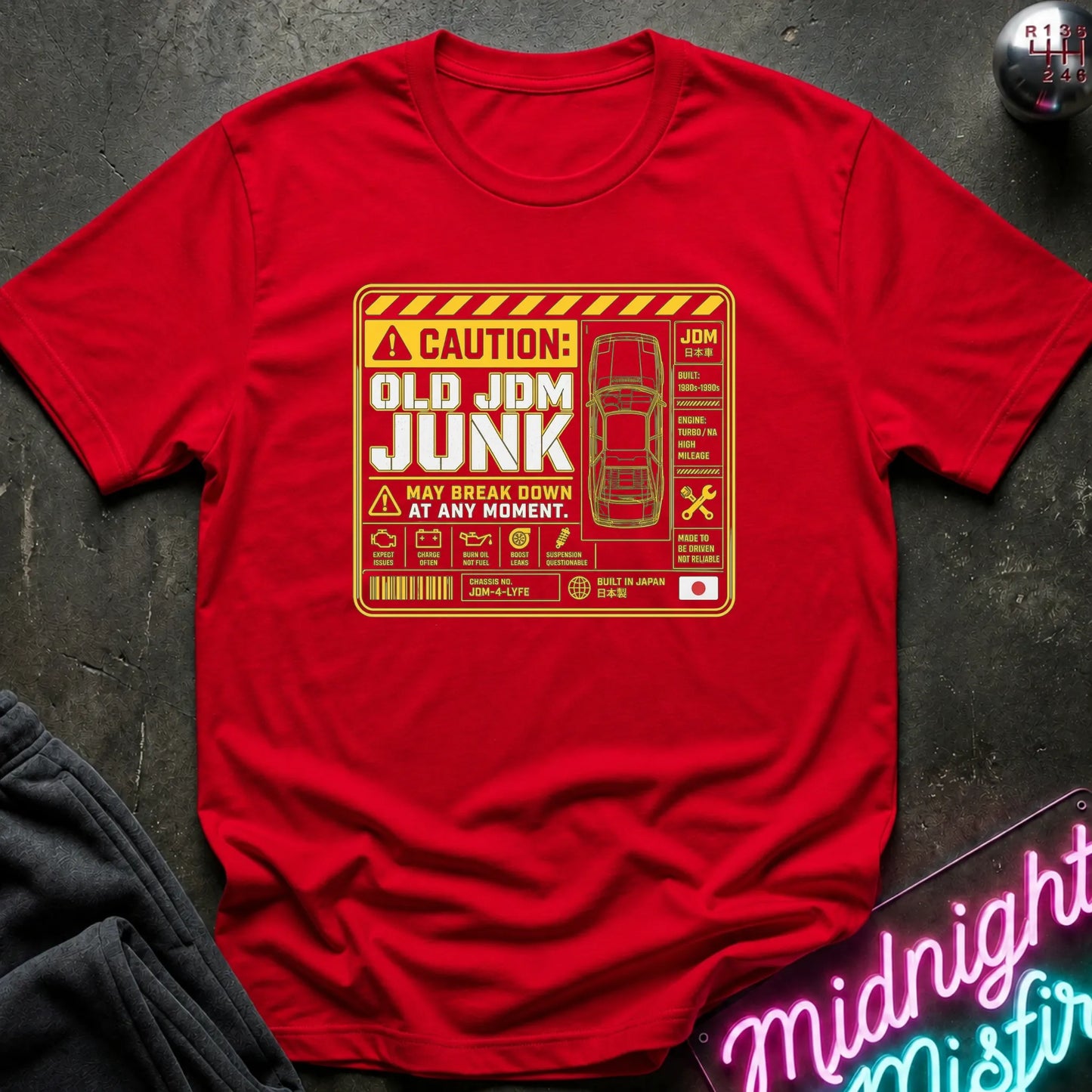 A red short-sleeve crewneck t-shirt with a yellow and white warning-style graphic on the chest reading 'CAUTION: OLD JDM JUNK MAY BREAK DOWN AT ANY MOMENT.' The shirt is laid flat on a dark textured surface with partial dark clothing and a neon sign visible at the edges.