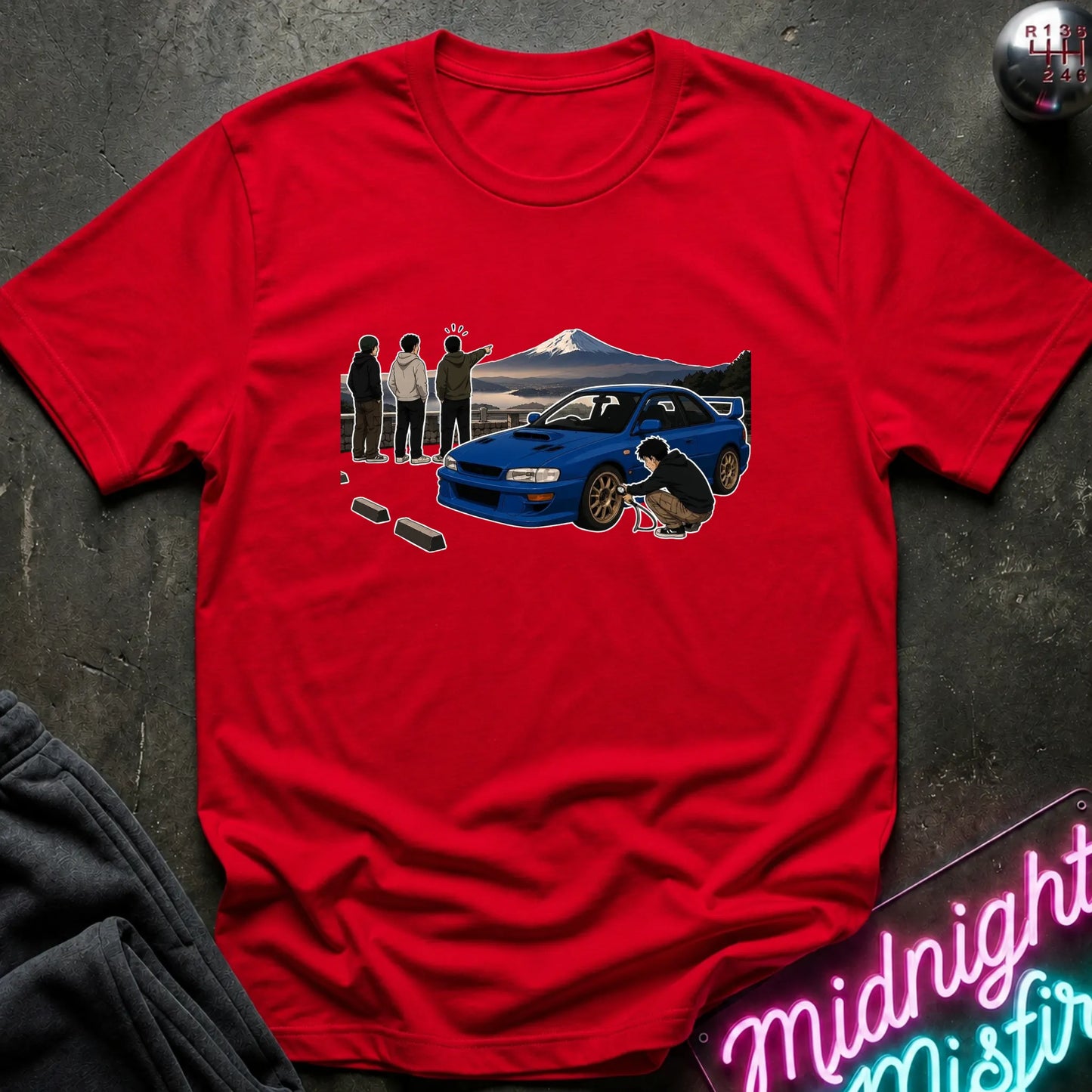A red short-sleeve crewneck t-shirt laid flat on a dark textured surface. The shirt features a graphic print of a blue Subaru WRX STI car, a mountain background, and three figures, with a neon sign and dark clothing partially visible in the styled flat-lay scene.