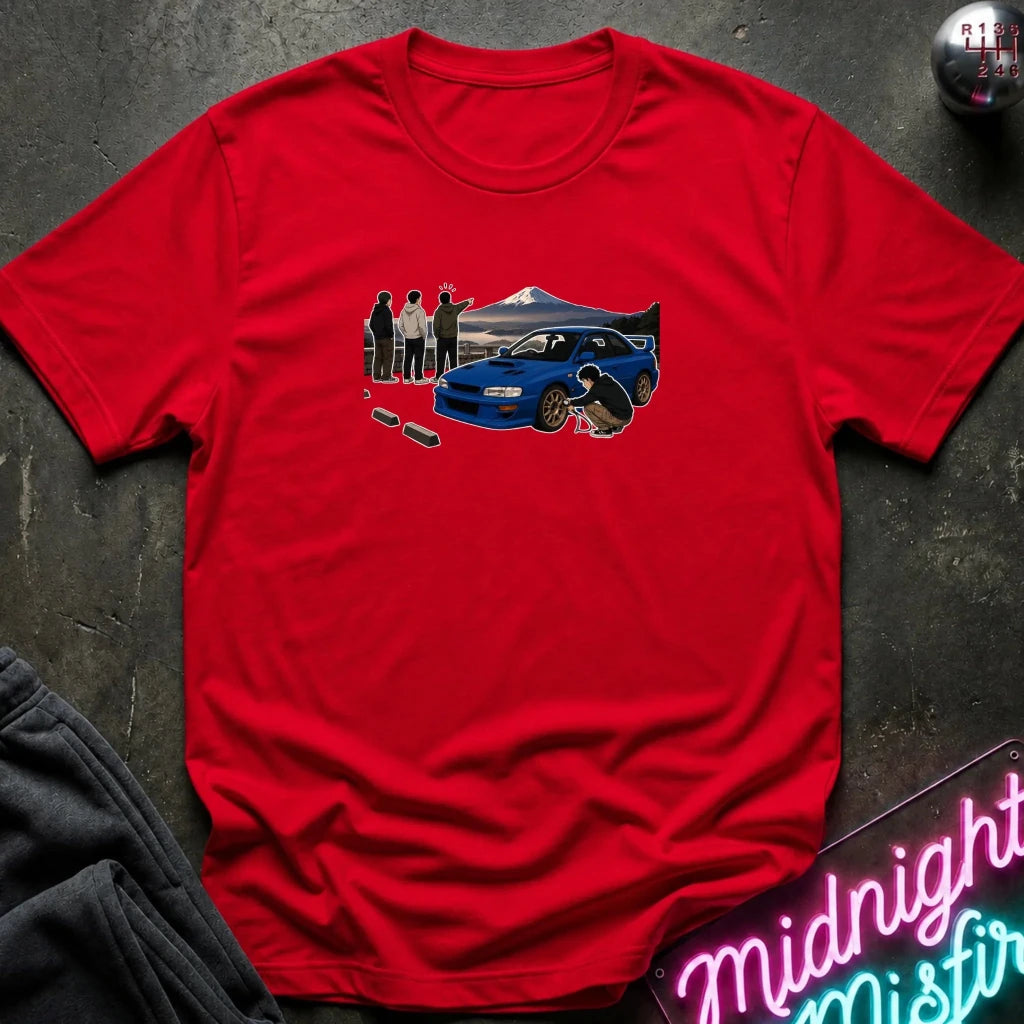 red crewneck t-shirt laid flat on a dark textured surface. The shirt features a graphic print of a blue sports car with a driver and spectators in front of a mountain landscape.