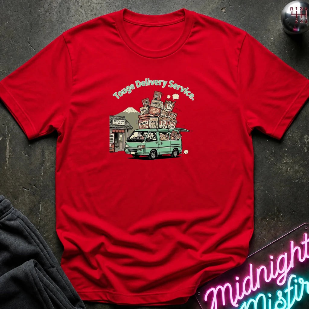 A red short-sleeve t-shirt laid flat on a dark textured surface, featuring a colorful graphic of a green van carrying stacked boxes with the text 'Touge Delivery Service.' printed above it.
