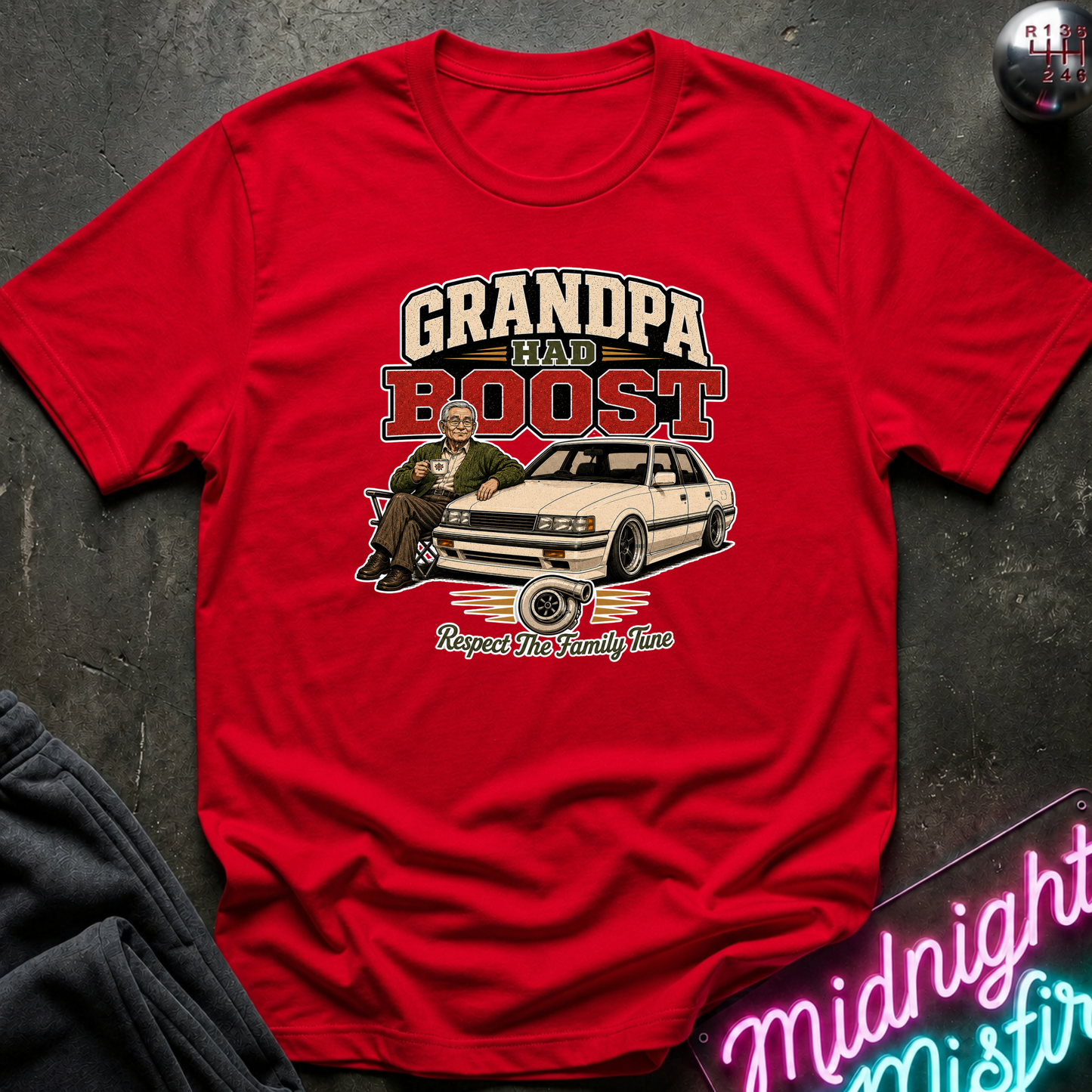 A red short-sleeve graphic T-shirt laid flat on a dark textured surface. The shirt features a large vintage-style print reading “GRANDPA HAD A BOOST” above an illustration of a man sitting in a white car, with smaller text below.