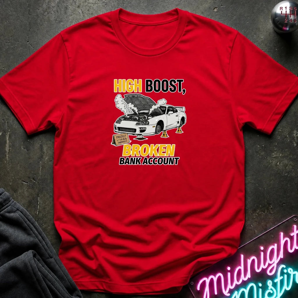 A red short-sleeve graphic T-shirt laid flat on a dark textured surface. The shirt features a printed design of a white car with its hood open, surrounded by smoke, with the text 'HIGH BOOST, BROKEN BANK ACCOUNT' in yellow and black.