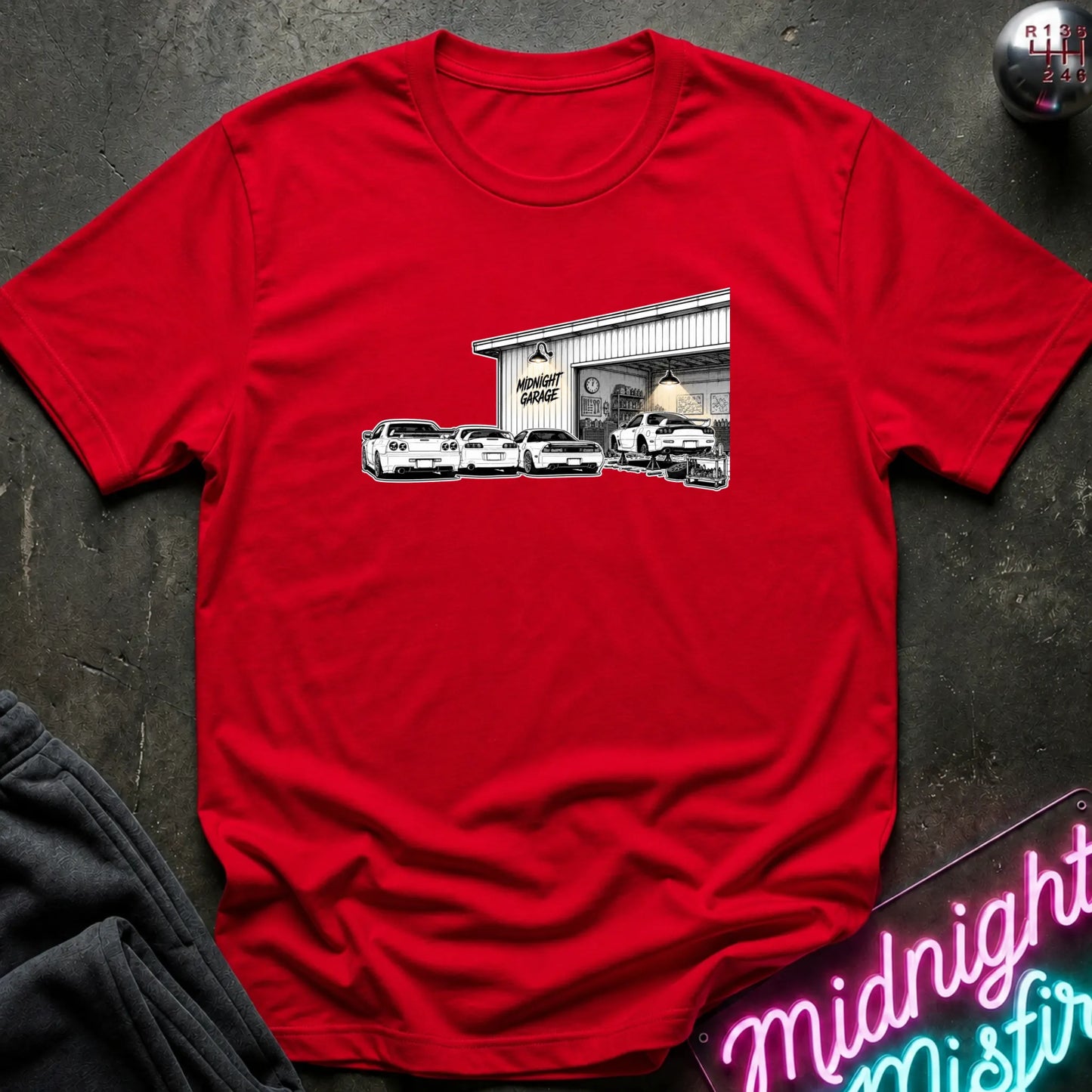 A red short-sleeve t-shirt laid flat on a dark textured surface, featuring a black and white graphic print of a garage with several cars in front. 