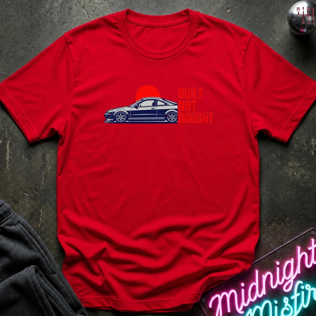 A red short-sleeve crewneck t-shirt laid flat on a dark textured surface. The shirt features a graphic of a blue sports car in front of a red sun with the orange text 'BUILT NOT BOUGHT' next to it.