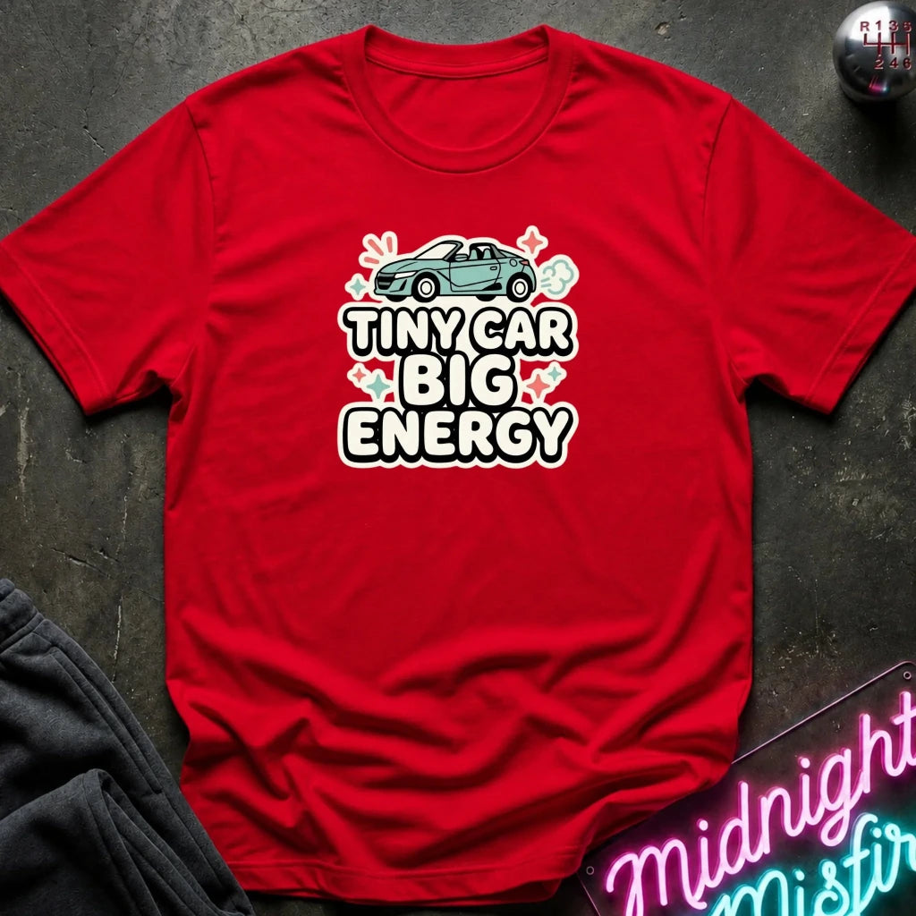 A red short-sleeve graphic T-shirt laid flat on a dark textured surface. The shirt features a cartoon car above the text 'TINY CAR BIG ENERGY' in white outlined letters with pink hearts.