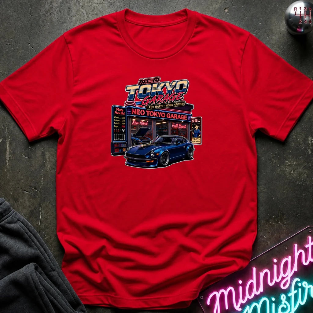 A red short-sleeve crewneck t-shirt featuring a 'Neo Tokyo Garage' graphic print with a blue sports car, laid flat on a dark textured surface with a neon sign and dark sweatpants partially visible.