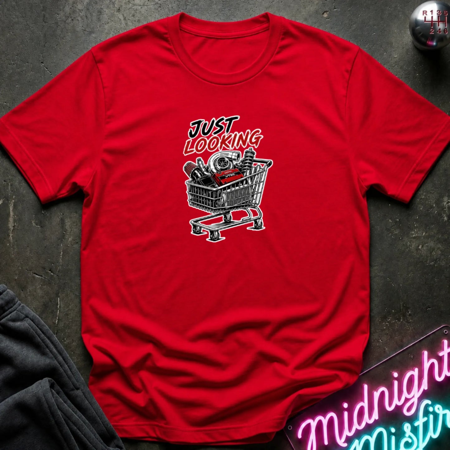 A red short-sleeve t-shirt laid flat on a dark textured surface, featuring a graphic print of a shopping cart filled with items and the text 'JUST LOOKING' in black and red. Partially visible props include dark pants in the lower left and a neon-style sign in the lower right.