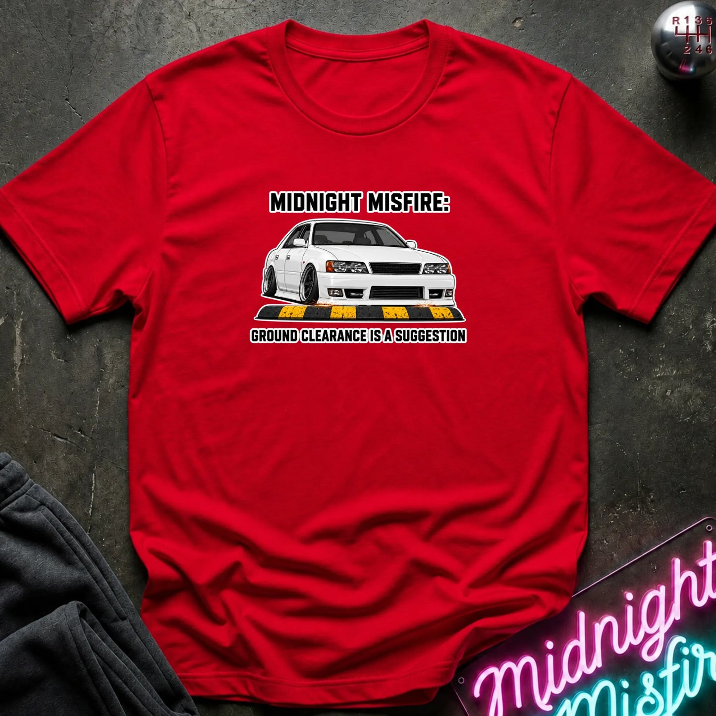 A red short-sleeve graphic t-shirt laid flat on a dark textured surface. The graphic features a white sports car with the text 'MIDNIGHT MISFIRE: GROUND CLEARANCE IS A SUGGESTION'.