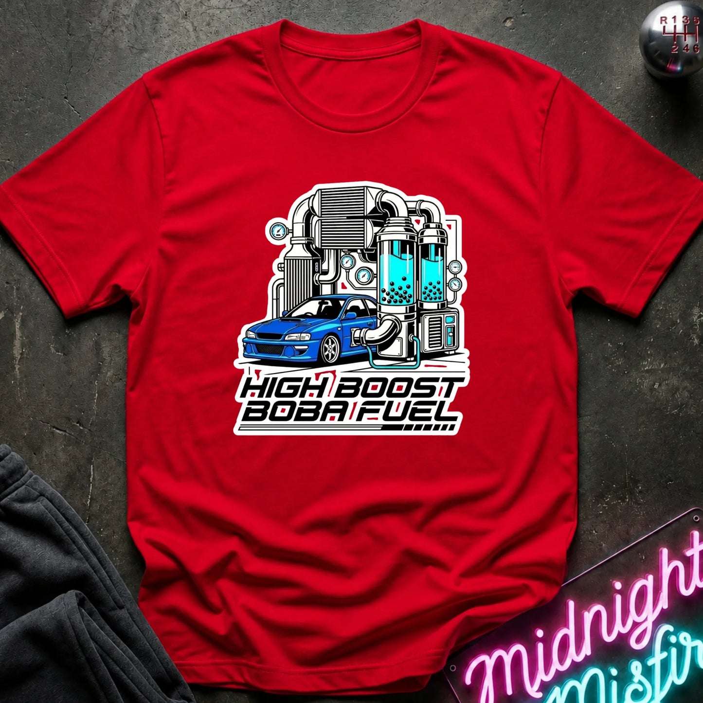 A red short-sleeve t-shirt laid flat on a dark textured surface, featuring a graphic of a blue car connected to a teal and gray high-performance turbo system with the text 'HIGH BOOST BOBA FUEL'.