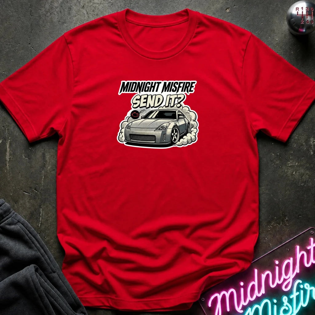A red short-sleeve graphic t-shirt laid flat on a dark textured surface. The graphic features the text 'MIDNIGHT MISFIRE SEND IT?' above a silver sports car with smoke effects.