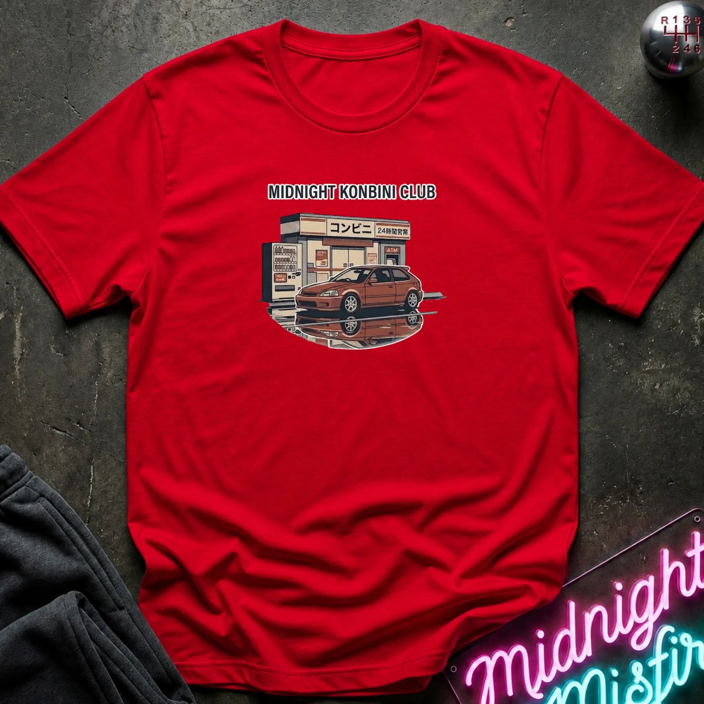A red short-sleeve graphic t-shirt laid flat on a dark textured surface. The graphic features a retro illustration of a car parked outside a convenience store with the text 'MIDNIGHT KONBINI CLUB' above it.