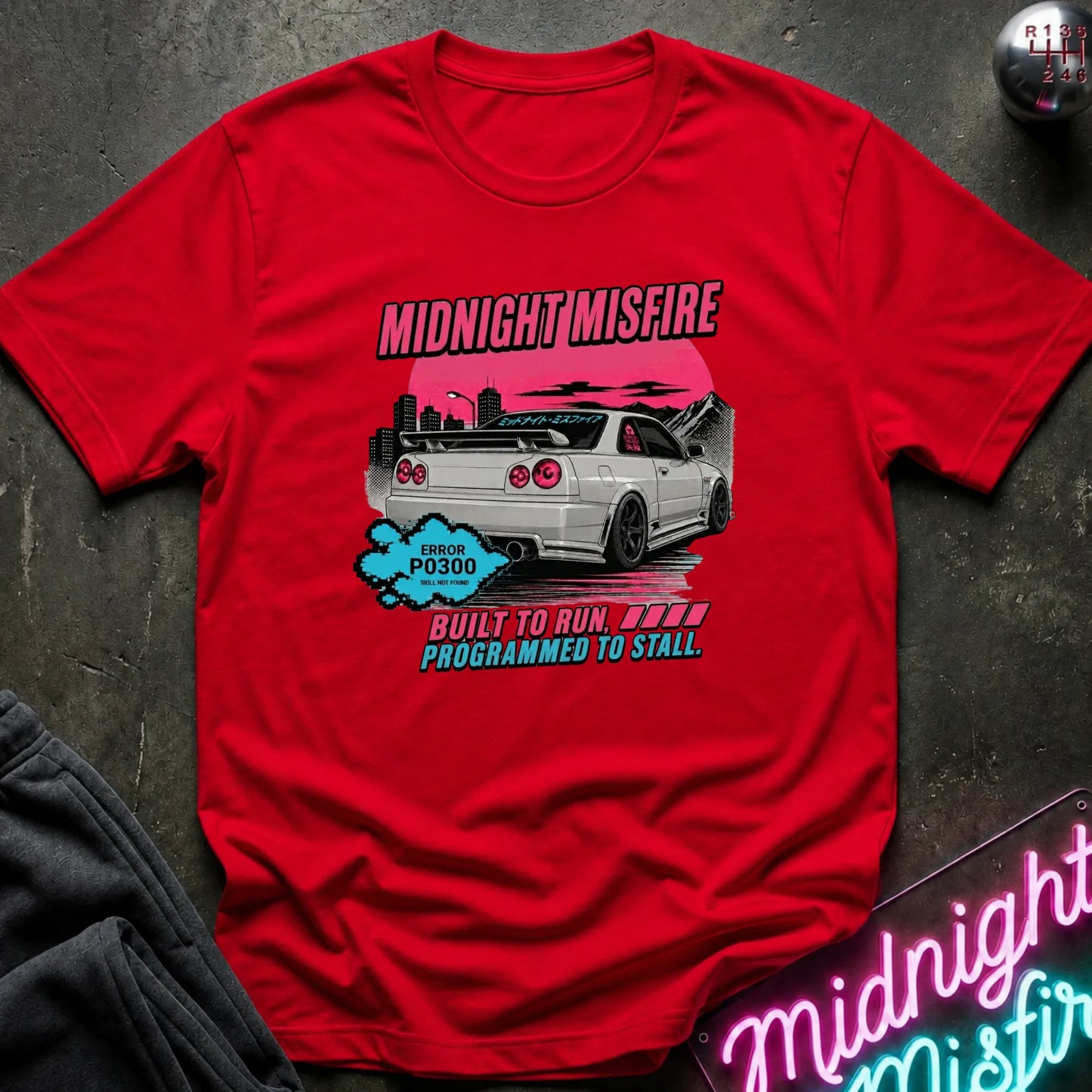 A red short-sleeve graphic t-shirt laid flat on a dark textured surface. The graphic features a white sports car with a pink sunset, the text 'MIDNIGHT MISFIRE' in pink.