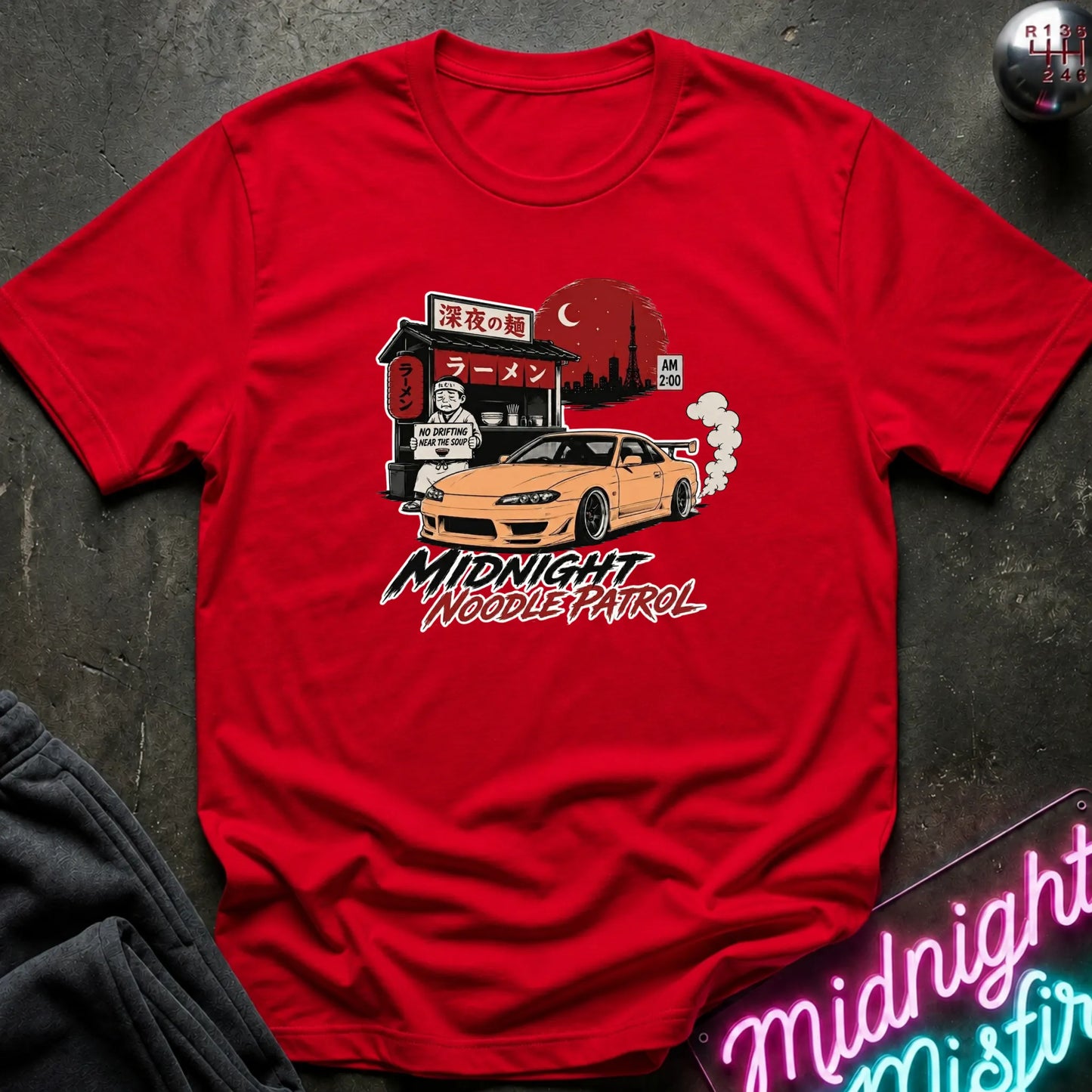 Red short-sleeve t-shirt laid flat on a dark textured surface, featuring a colorful graphic print of an orange sports car in front of a ramen shop at night with the text 'Midnight Noodle Patrol'.
