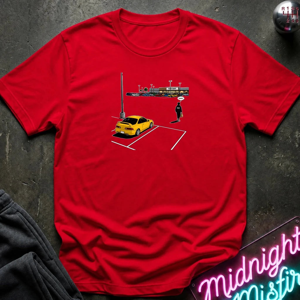 A red short-sleeve t-shirt laid flat on a dark textured surface, featuring a small graphic print of a yellow car, a parking lot, and a lone figure. 