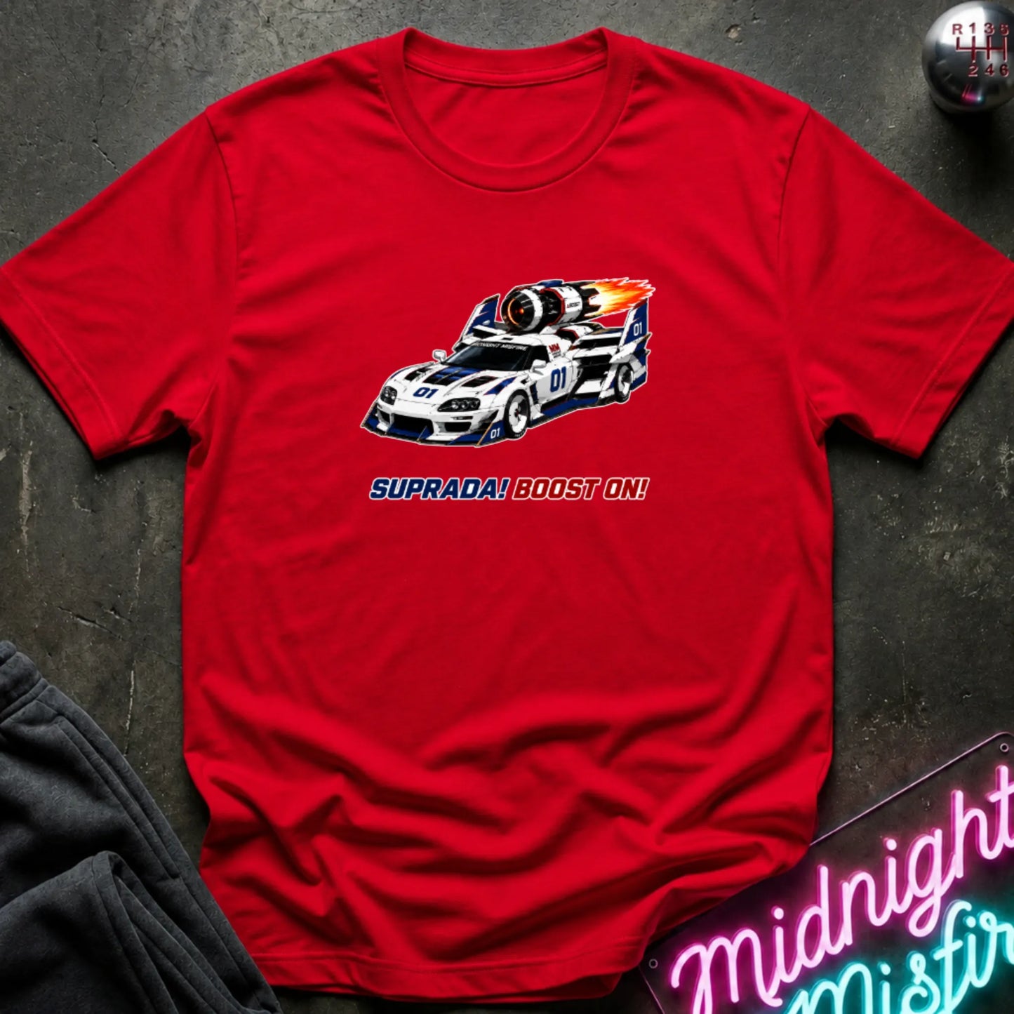 A red short-sleeve t-shirt with a retro-style graphic of a race machine with big boost, displayed flat on a dark textured surface with a neon sign and sweatpants partially visible.