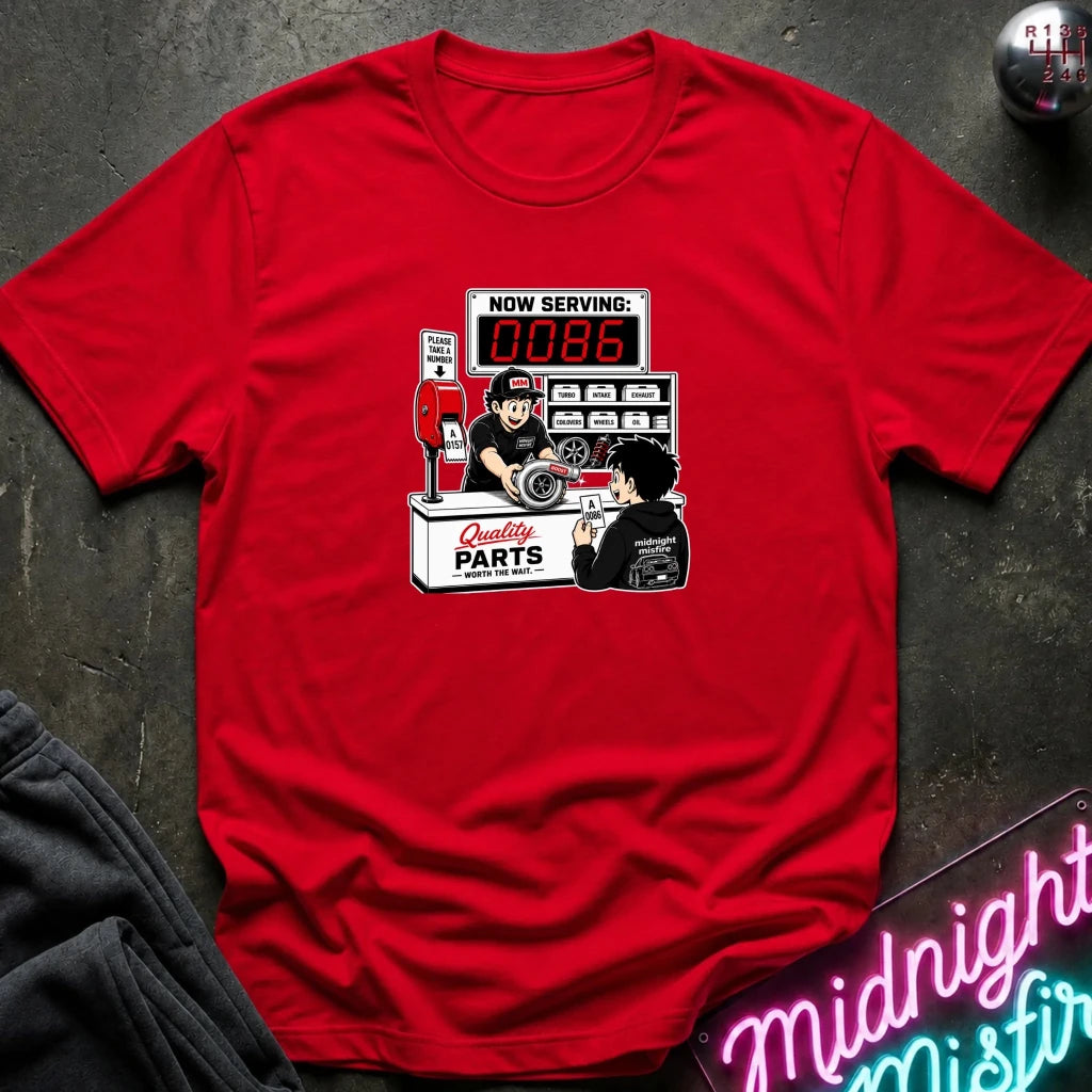 A red short-sleeve crewneck t-shirt featuring a retro-style graphic of a mechanic working at a parts counter with a digital display showing 'NOW SERVING: 0086'. 