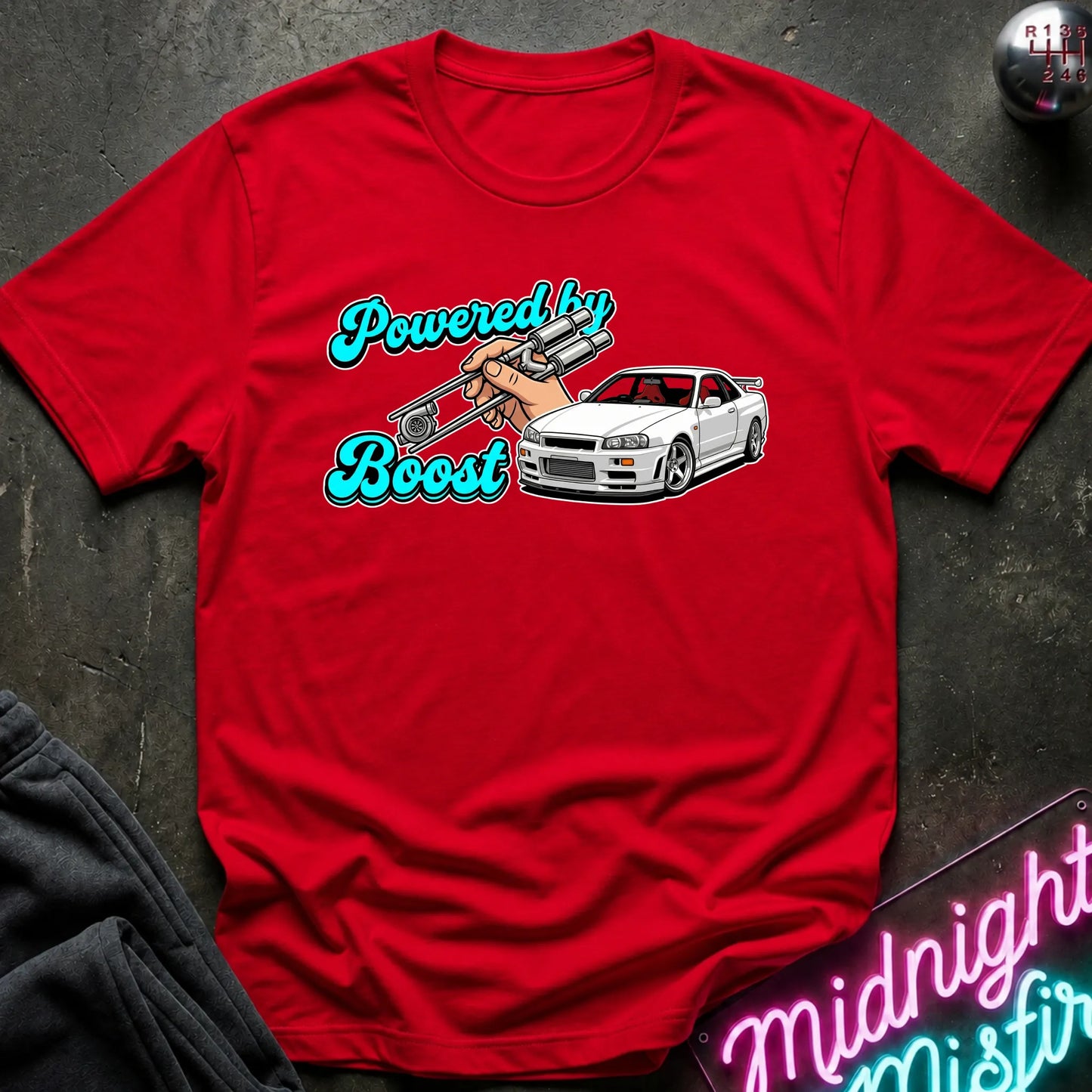 A red short-sleeve graphic T-shirt laid flat on a dark textured surface. The shirt features a white car illustration and turquoise text reading 'Powered by Boost.