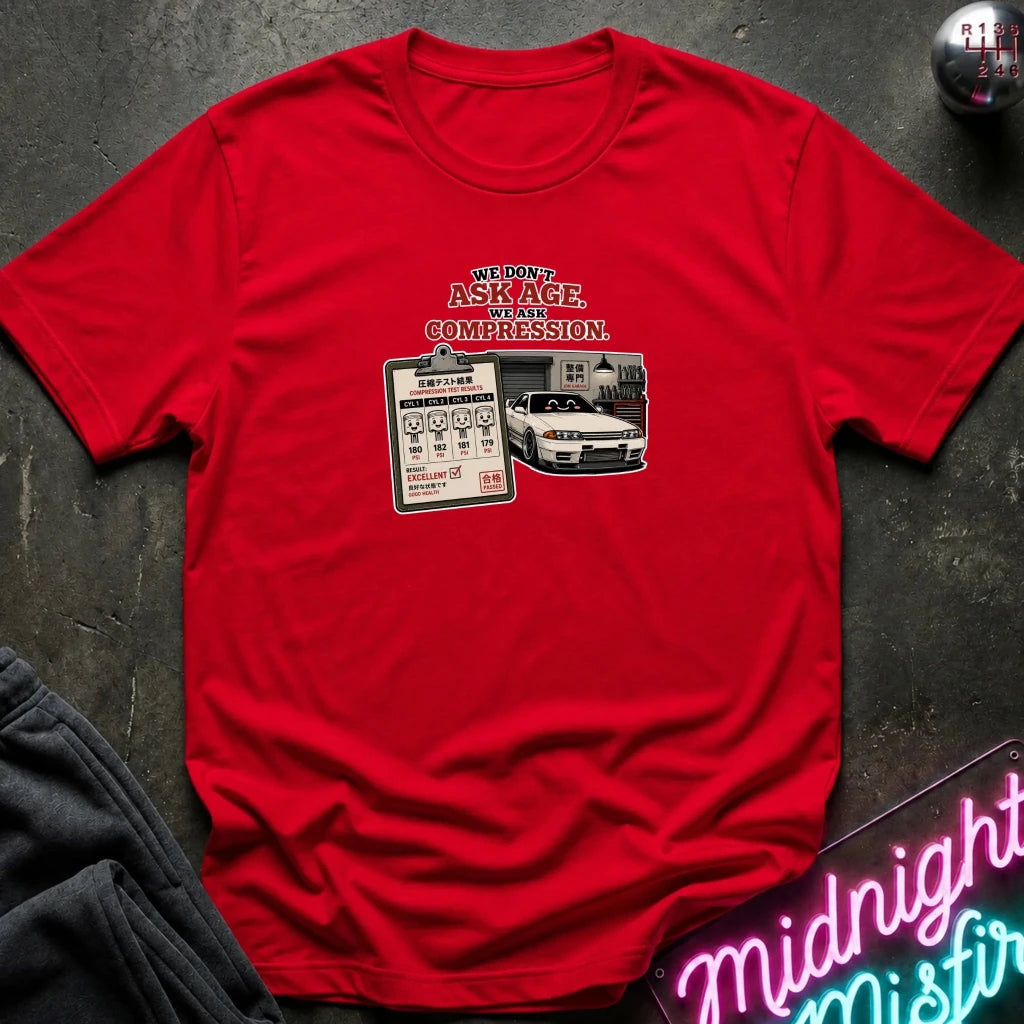 A red short-sleeve graphic t-shirt laid flat on a dark textured surface. The graphic features the text 'WE DON'T ASK AGE, WE ASK COMPRESSION.