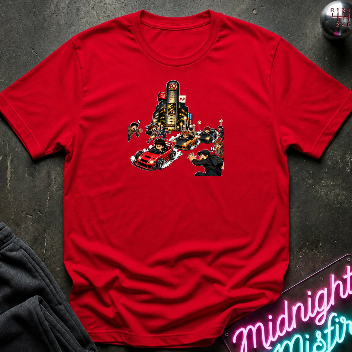 A red short-sleeve crewneck t-shirt with a graphic print of a city street scene featuring a red sports car and small figures, laid flat on a dark textured surface with a neon sign and dark pants partially visible.