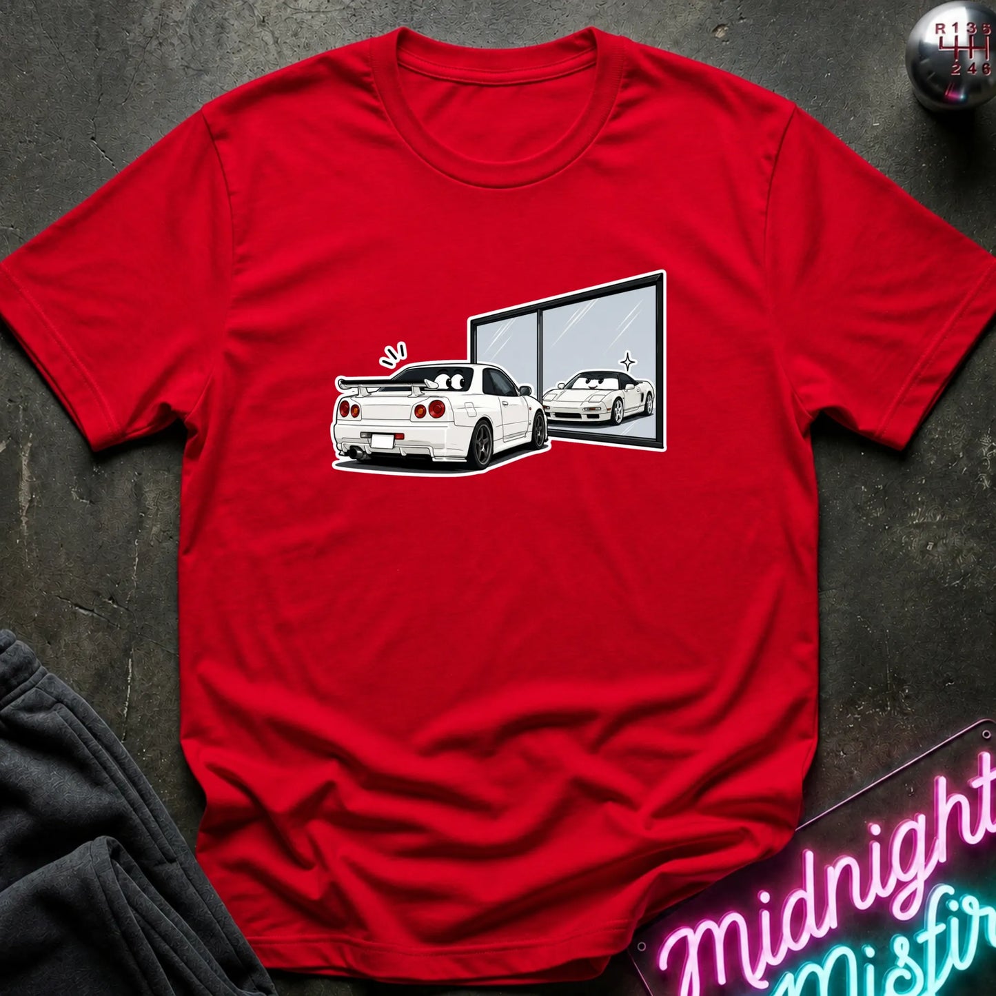 A red short-sleeve crewneck t-shirt featuring a graphic print of a white sports car and its reflection in a window, laid flat on a dark textured surface with a neon sign and dark sweatpants partially visible.